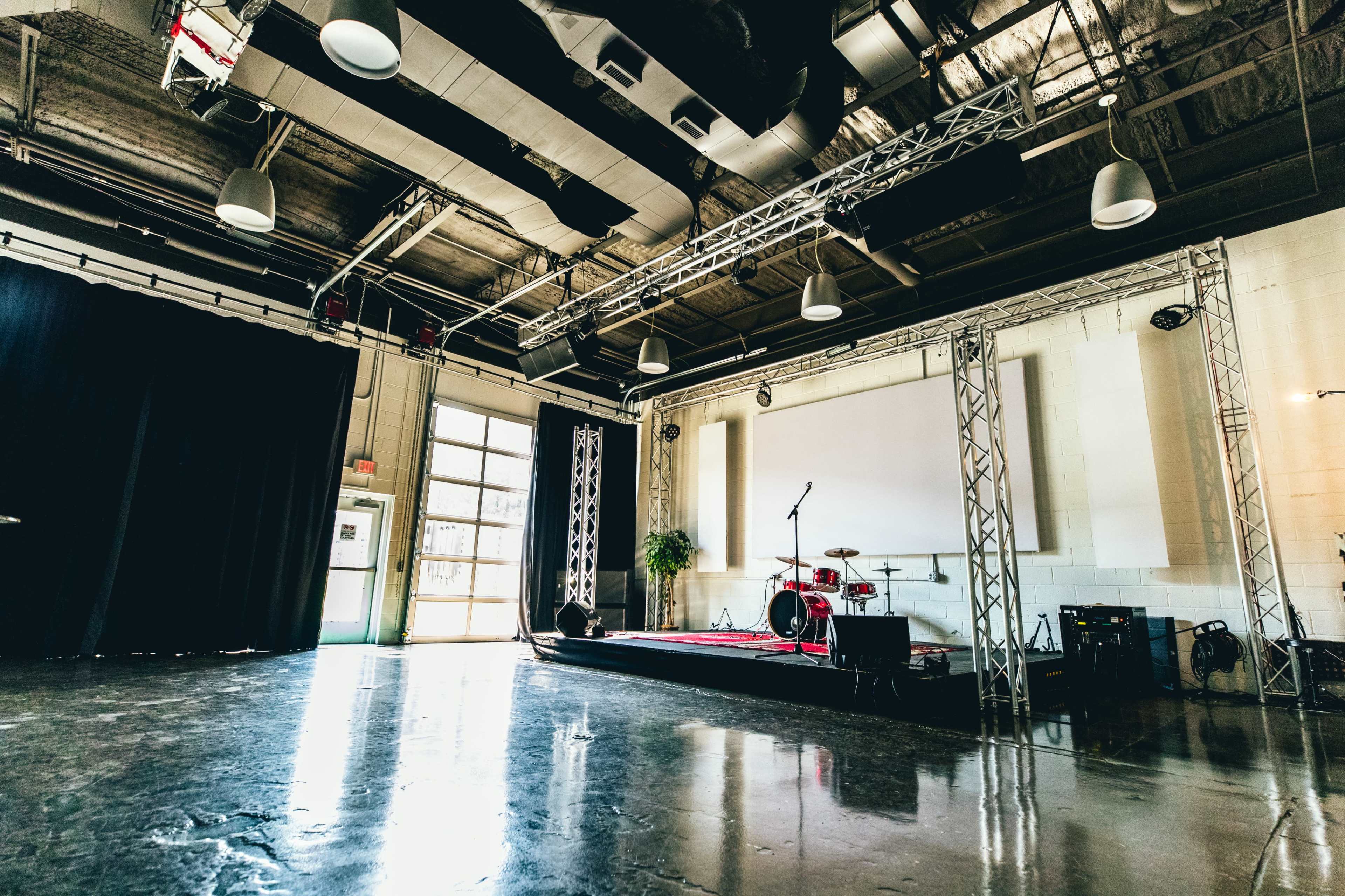 The image shows an empty performance space with a small stage, drum set, and black curtains, illuminated by overhead lights.
