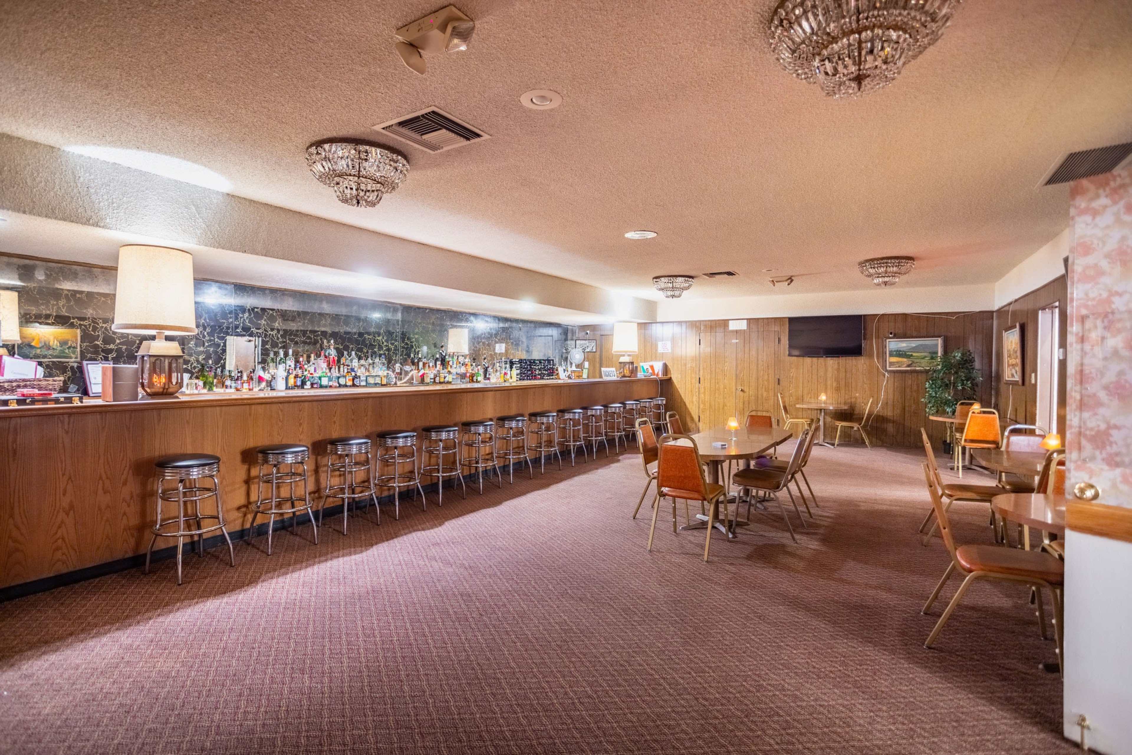 The image shows a spacious bar area with a long counter, several high-backed stools, and tables arranged in the foreground, all under soft lighting and decorative chandeliers.