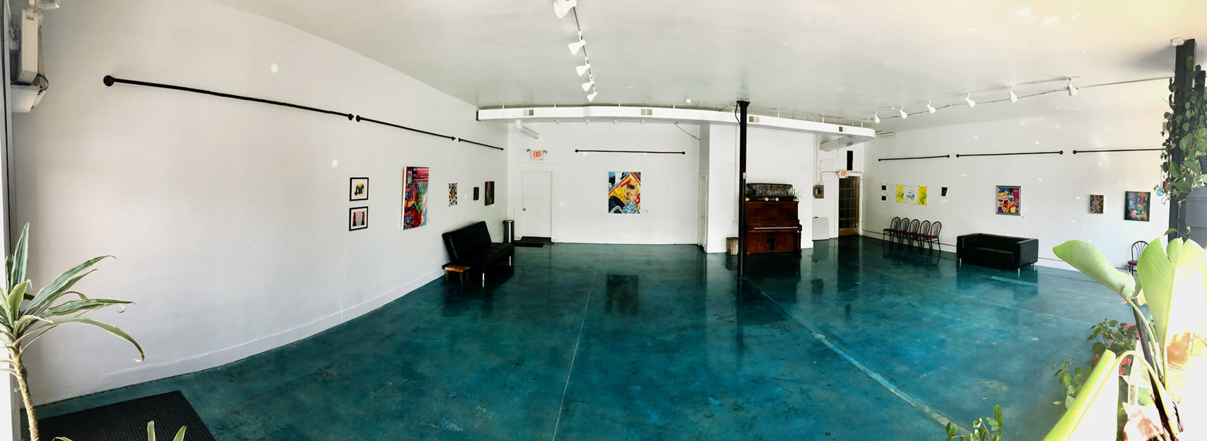 The image shows a spacious, minimalist gallery with a polished blue floor, black seating, and several artworks displayed on the white walls.