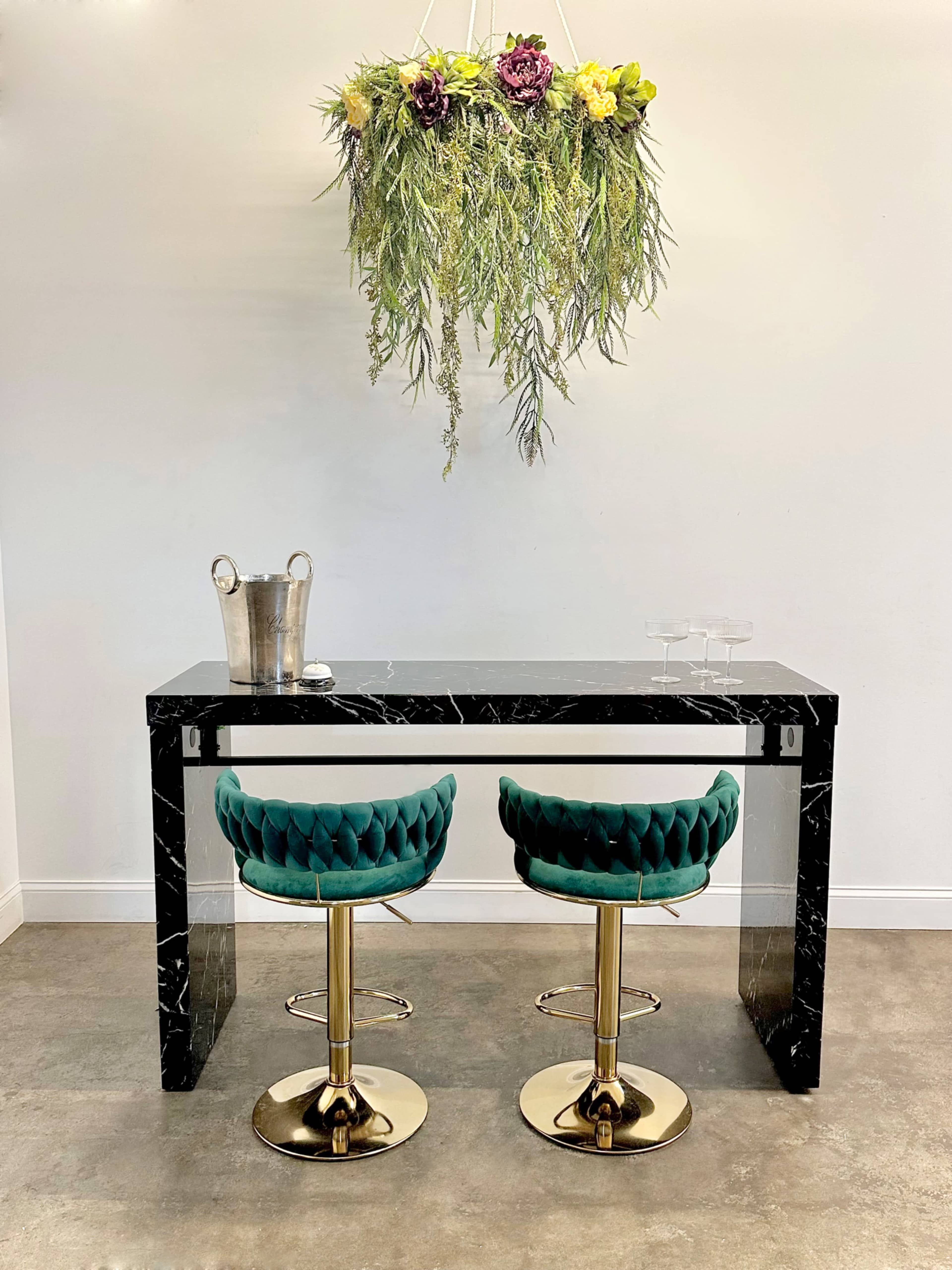 A modern black marble table with two green velvet barstools sits beneath a hanging floral arrangement.