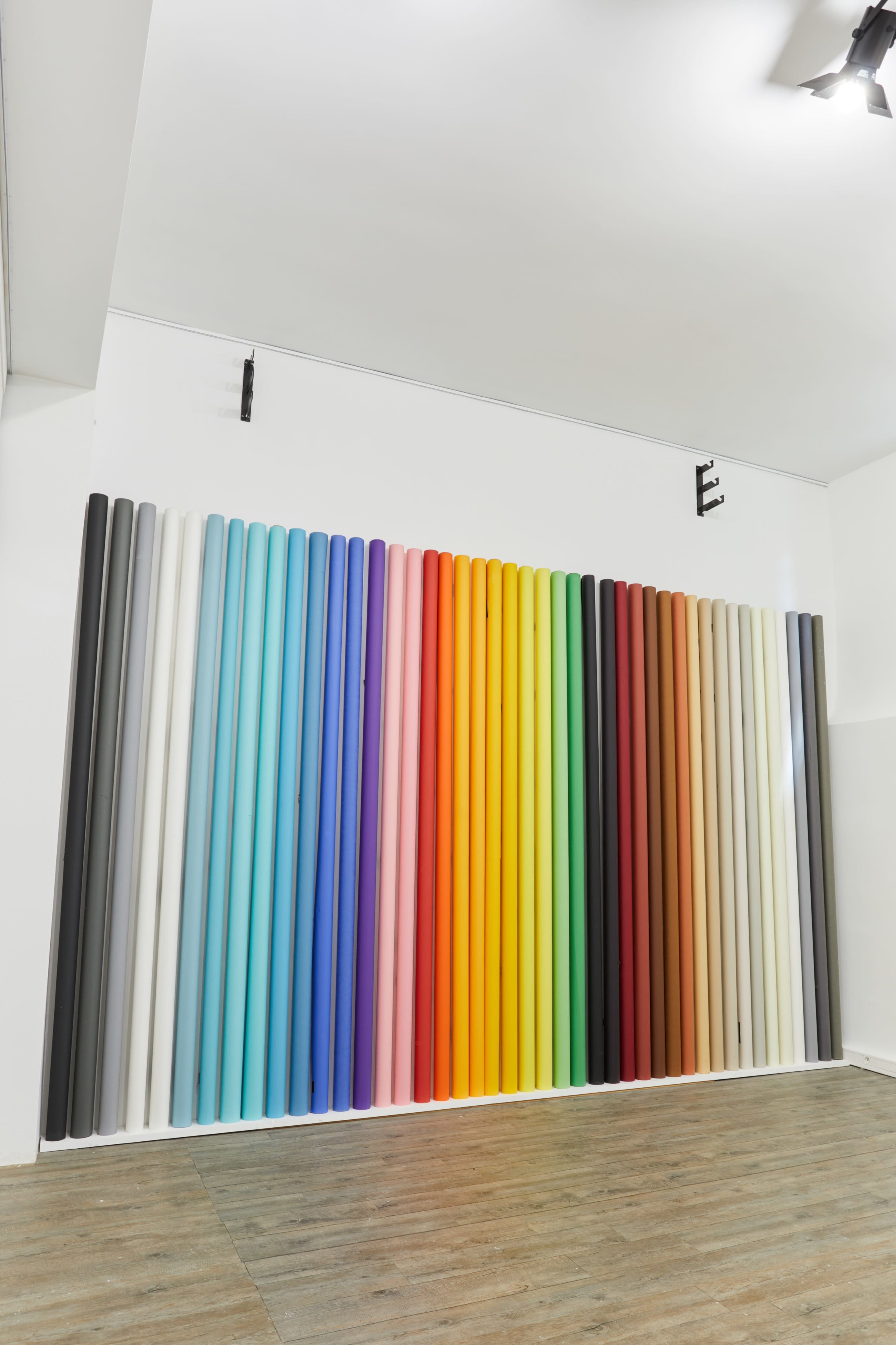The image features a wall covered with vertical rolls of colored paper arranged in a gradient from dark to light shades.