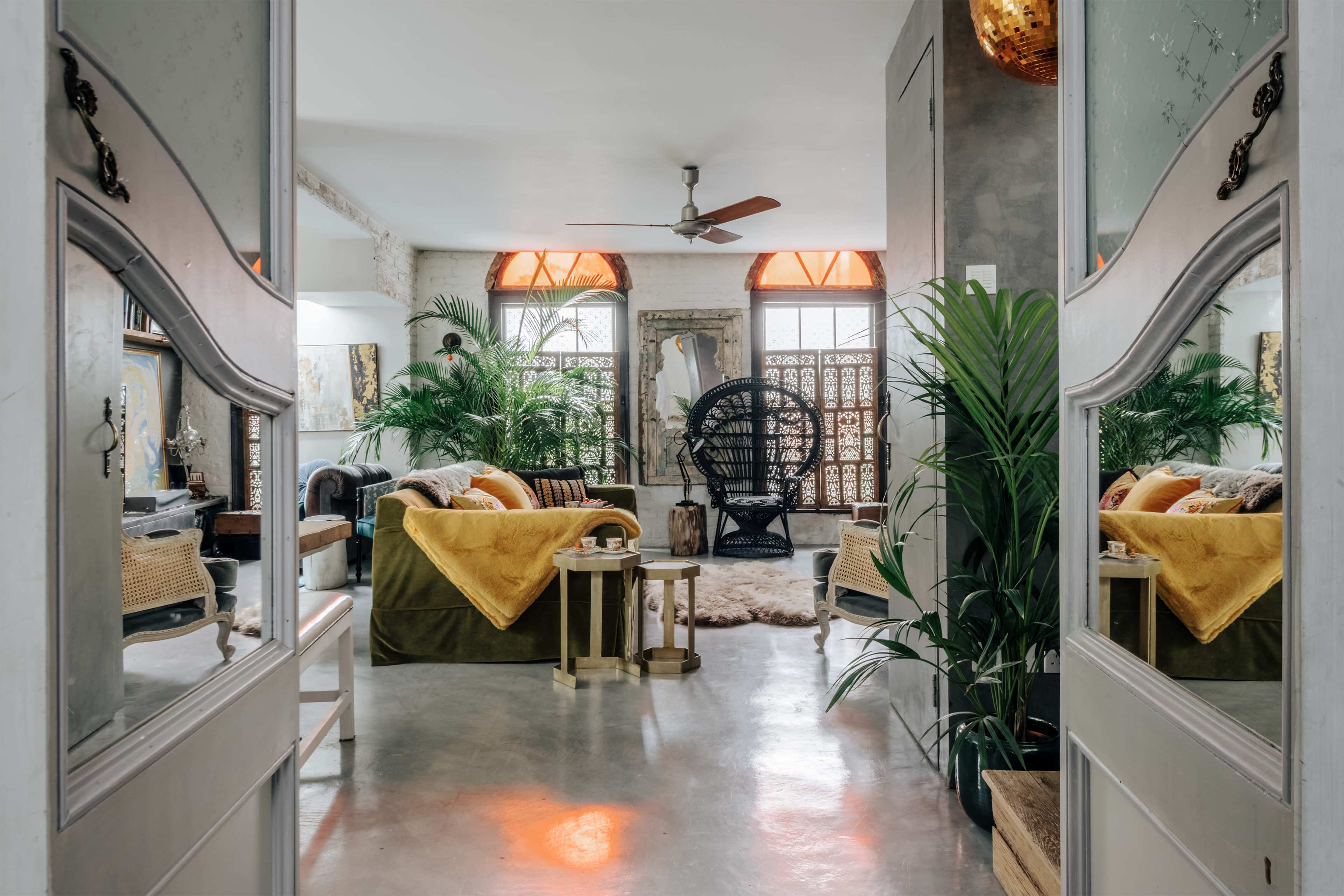 The image shows a spacious living room with a mix of modern and bohemian decor, featuring a large green sofa, a hanging chair, and numerous houseplants.