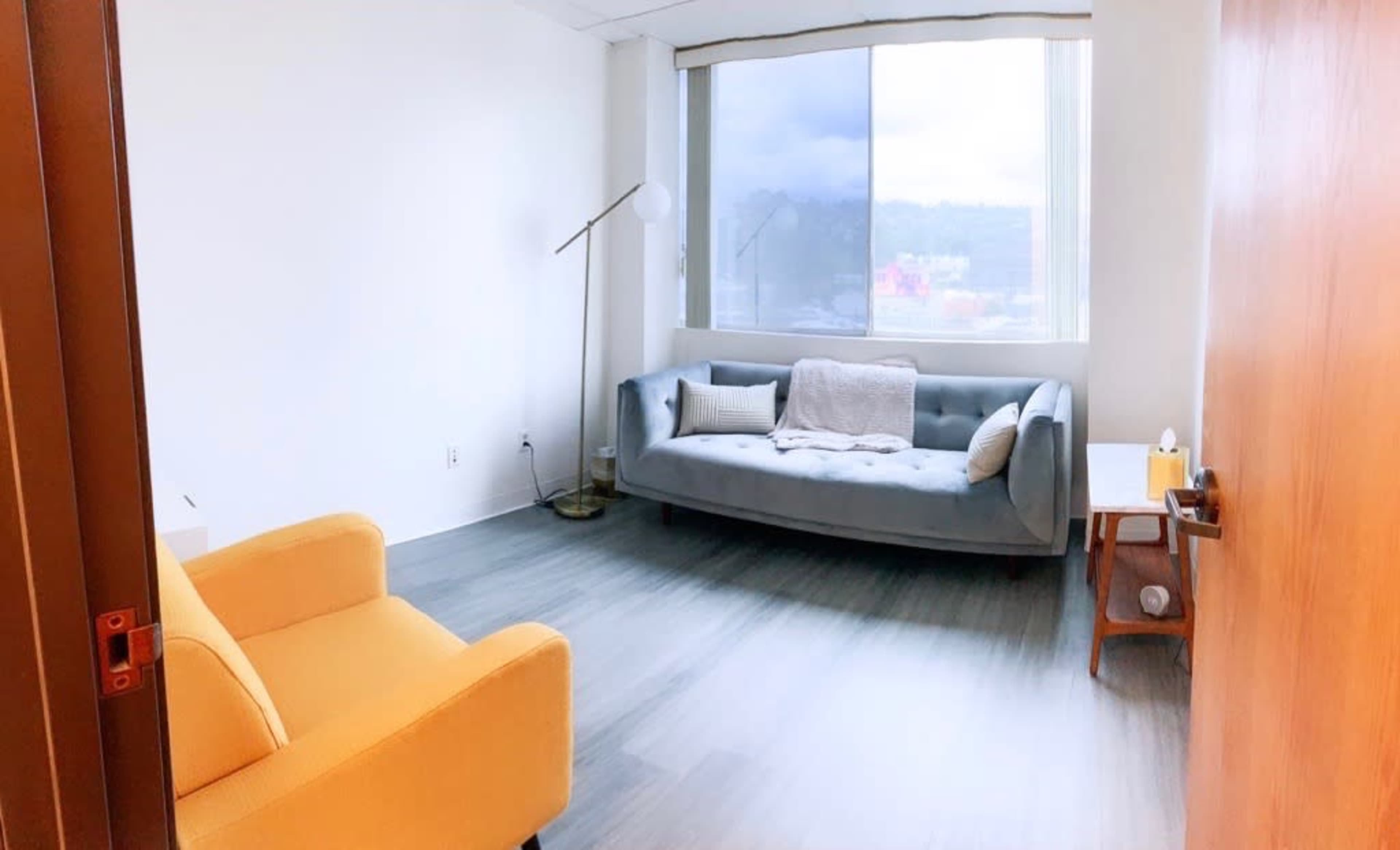 A light-colored room with a blue sofa, a yellow armchair, a floor lamp, and a small side table near a large window.