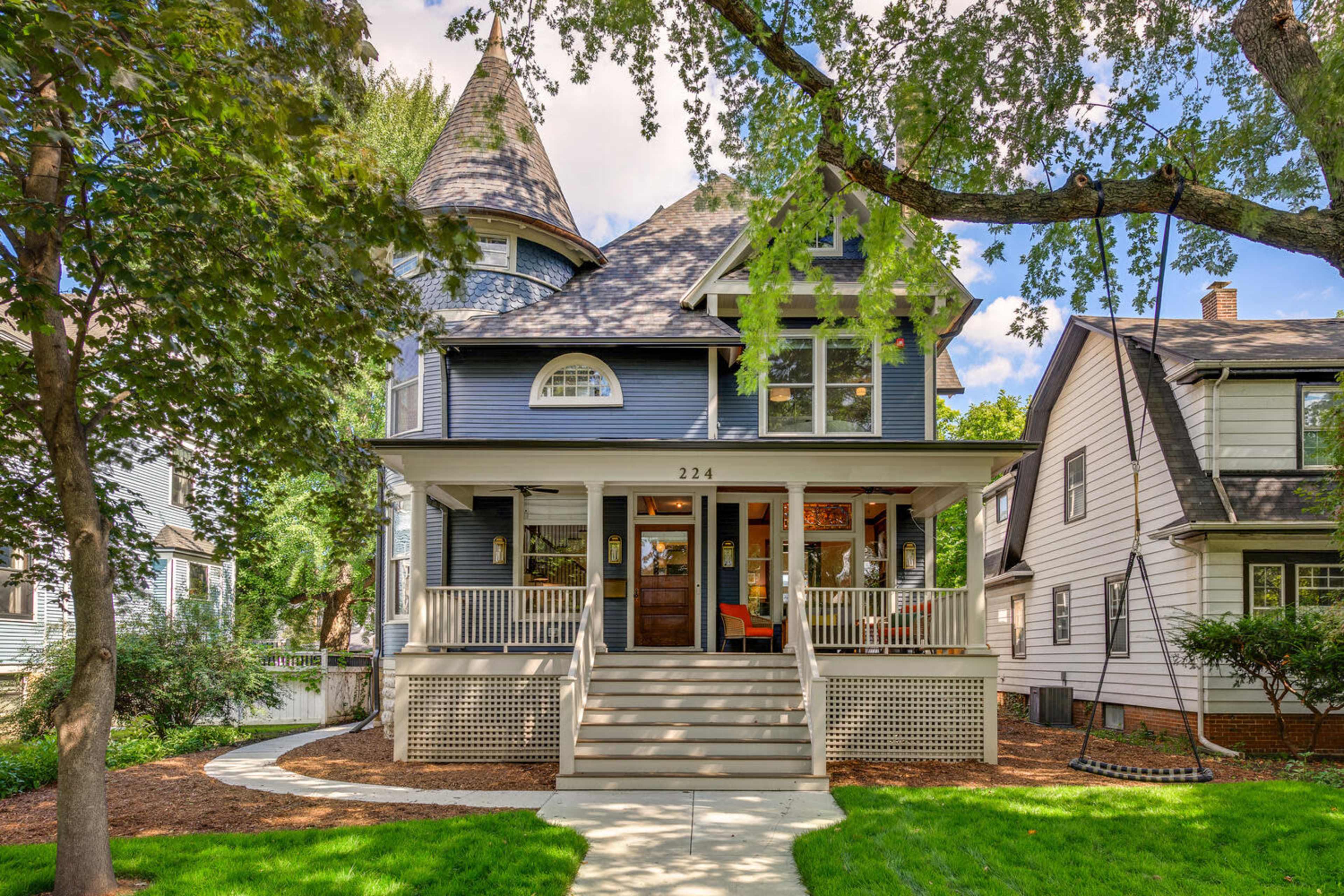 A large, multi-story blue house with a turret and a welcoming front porch is surrounded by trees and lawns.