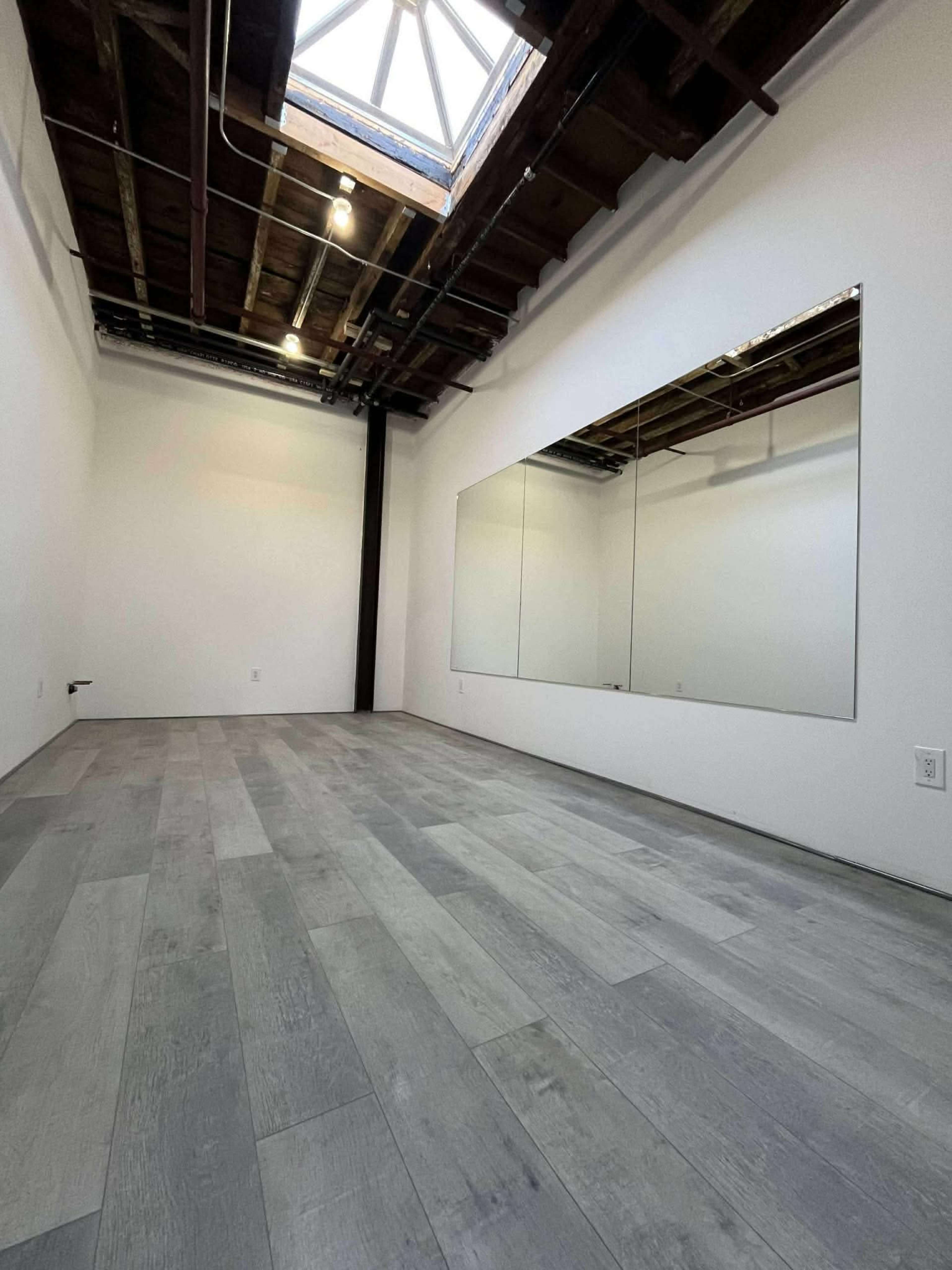 The image depicts a spacious, empty room with wooden floors, a skylight, and a large mirror on one wall.