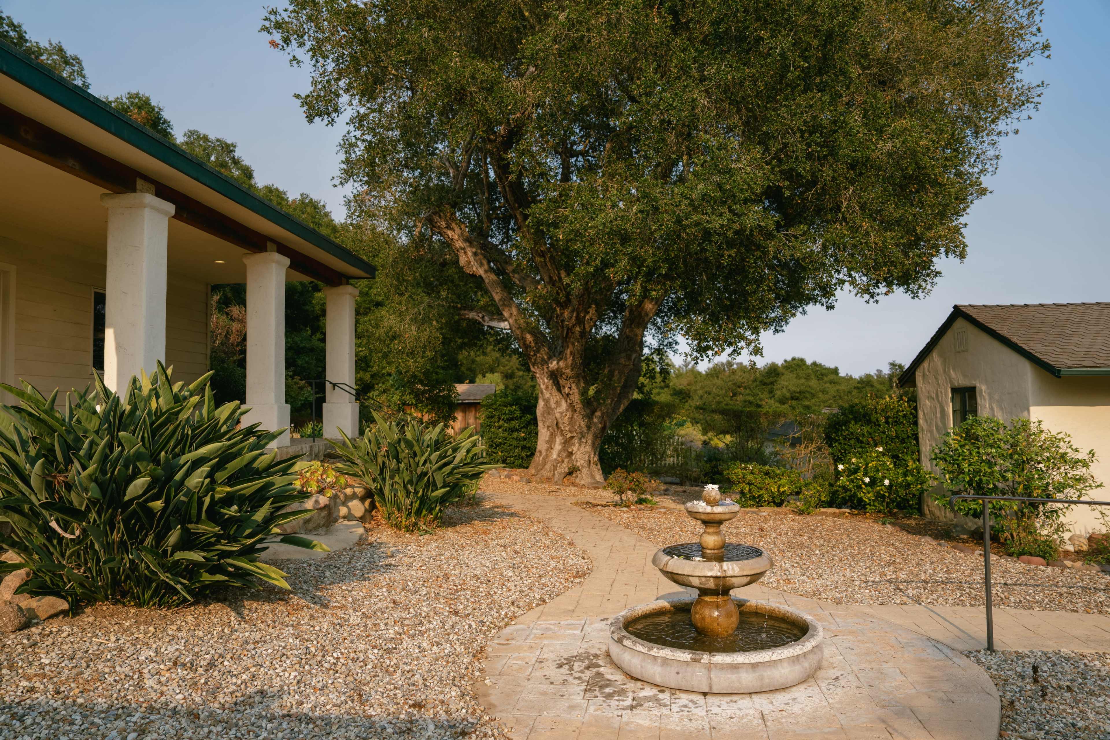 A stone pathway leads to a three-tiered fountain surrounded by gravel and greenery, with a large tree nearby and a house in the background.