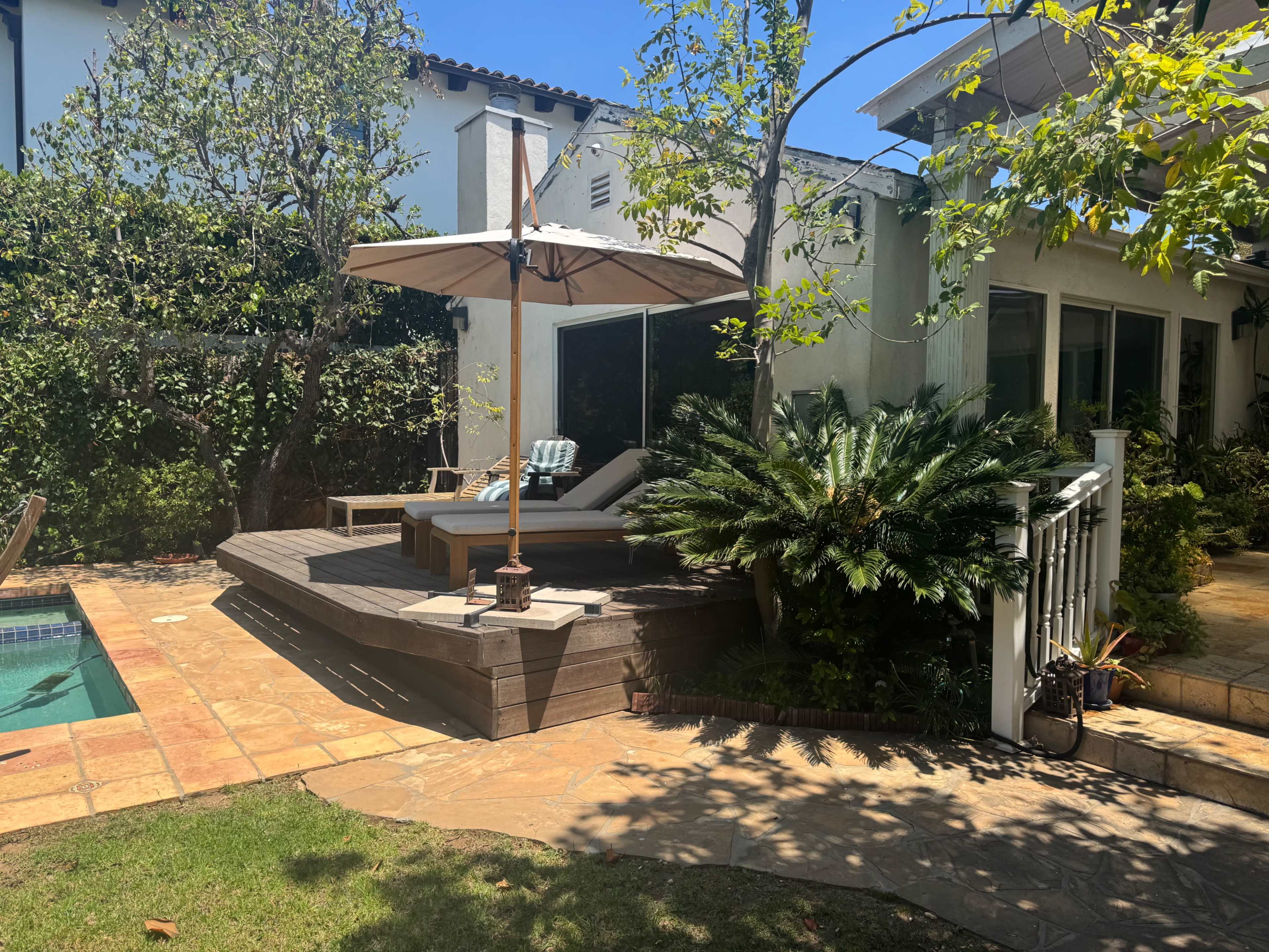 The image shows a wooden deck with a lounge chair and an umbrella, surrounded by greenery near a swimming pool and a house.