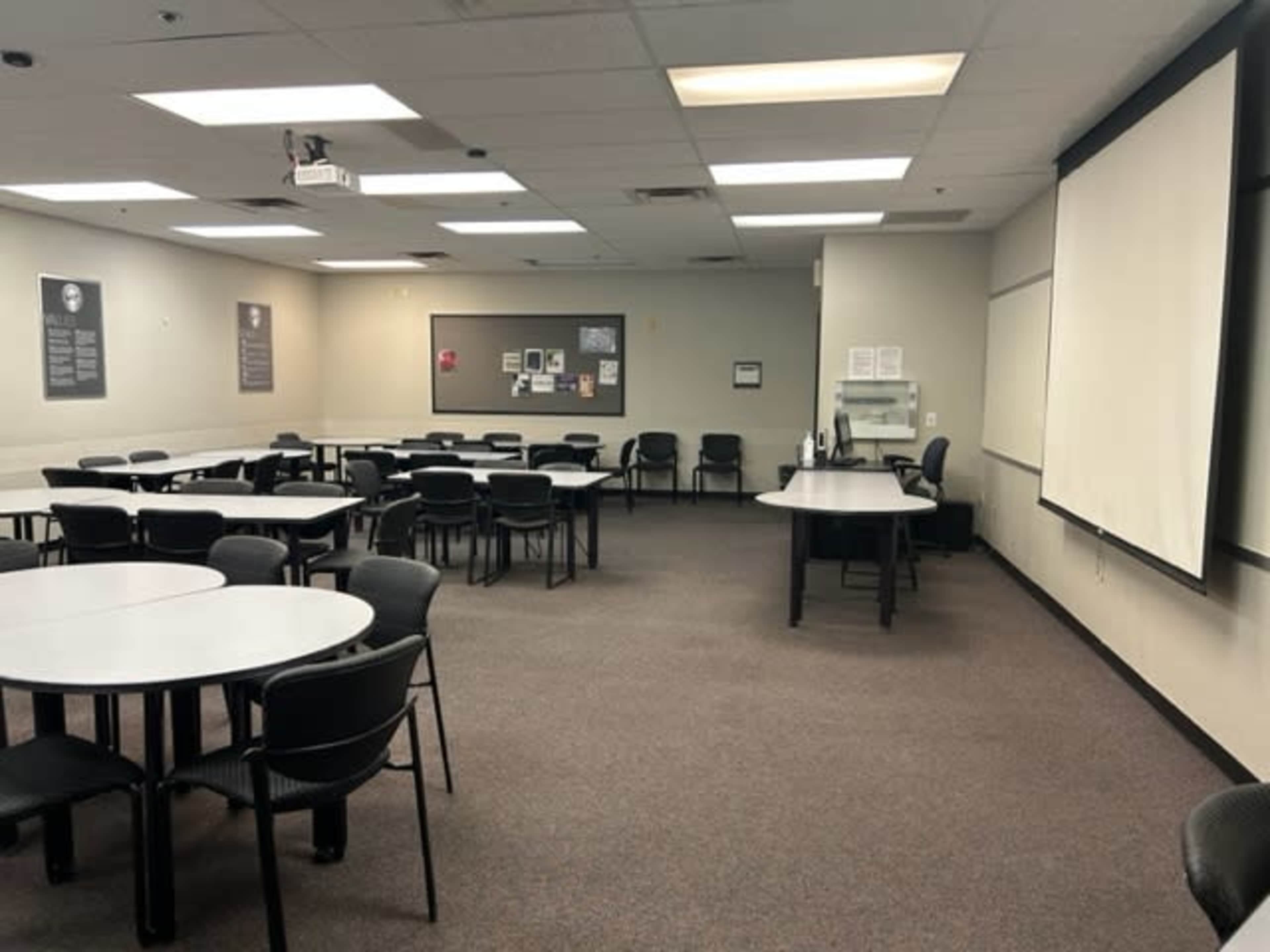 A classroom with rows of circular tables and chairs, a projector, and a whiteboard at the front.