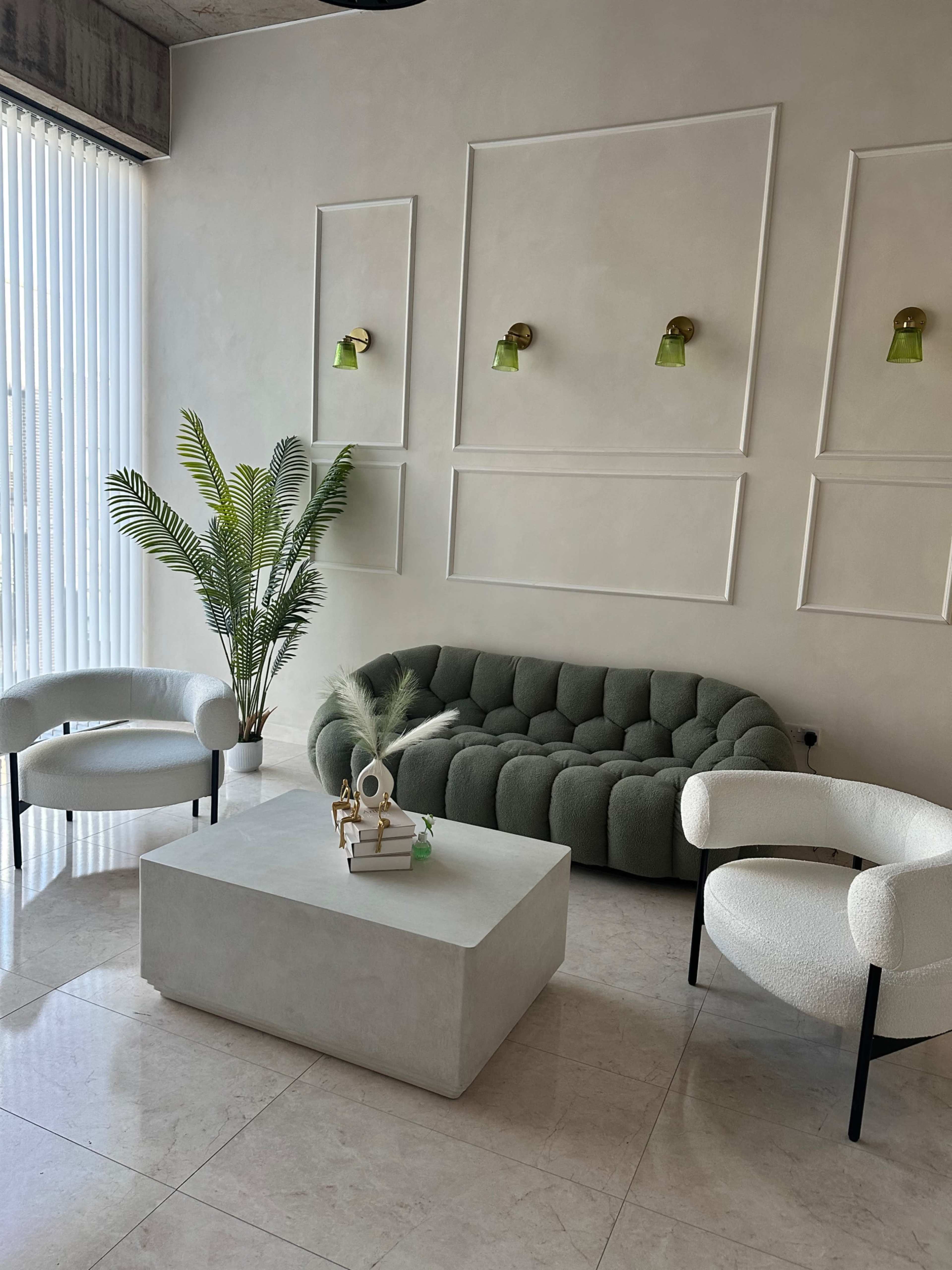 The image showcases a modern living space with a gray, tufted sofa, two white armchairs, a light gray coffee table, and a tall indoor plant, all set against a neutral wall with green wall sconces.