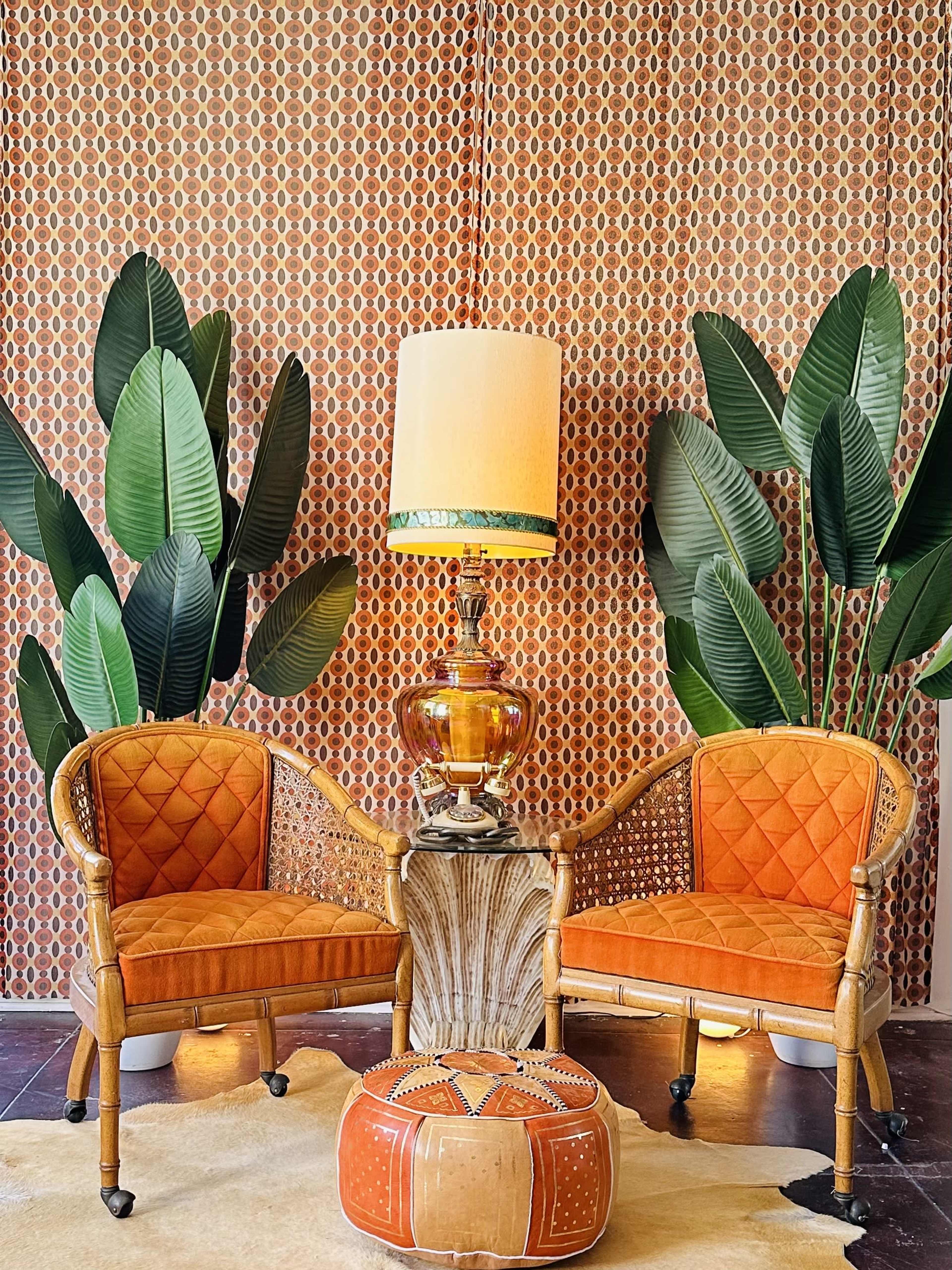 Two orange velvet chairs are positioned on either side of a decorative table, with a large lamp and tall green plants in the background against a patterned wall.