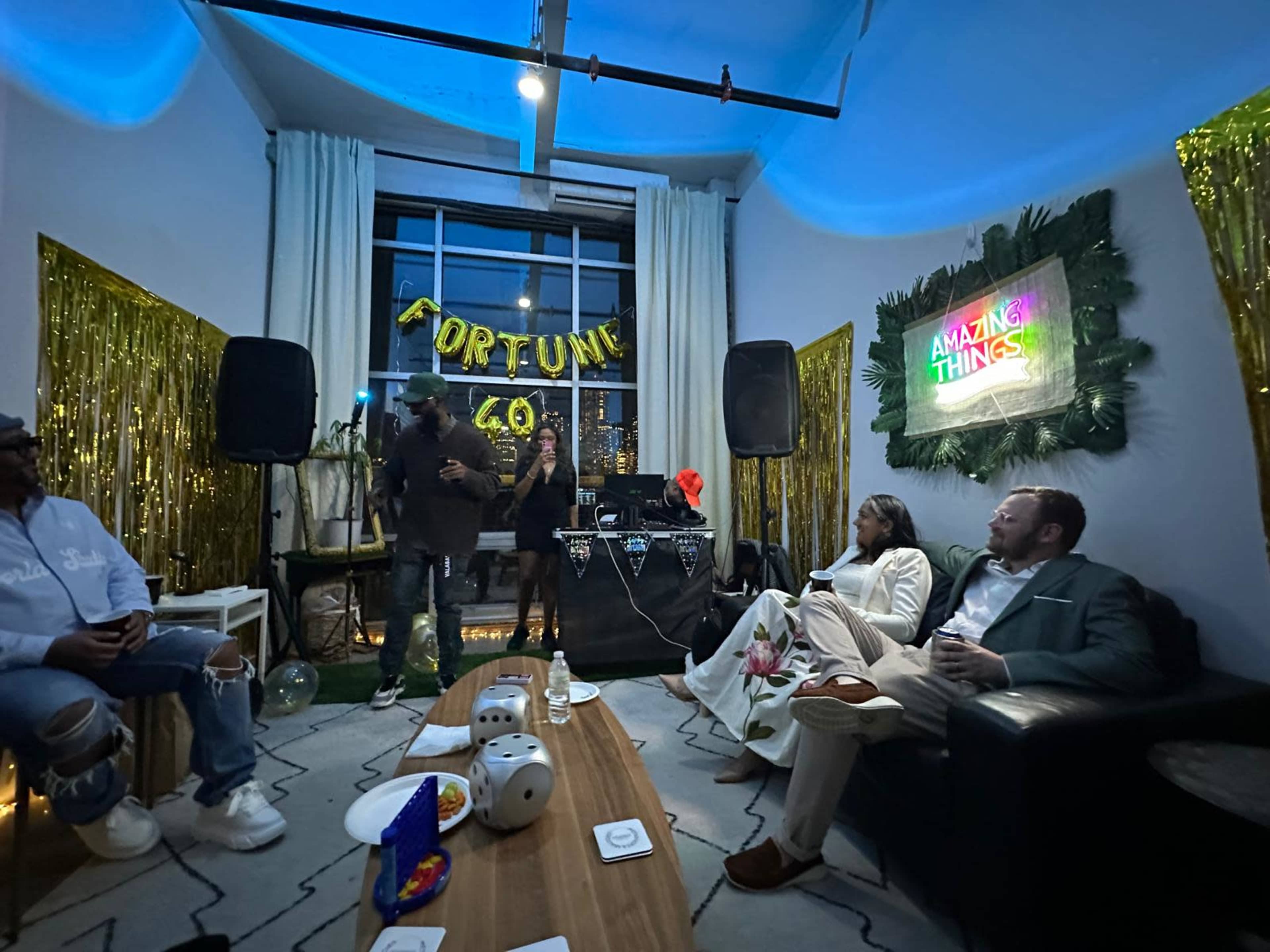 The image depicts a festive indoor gathering with a DJ setup, decorations for a 60th birthday party, and guests sitting on a sofa and chairs.