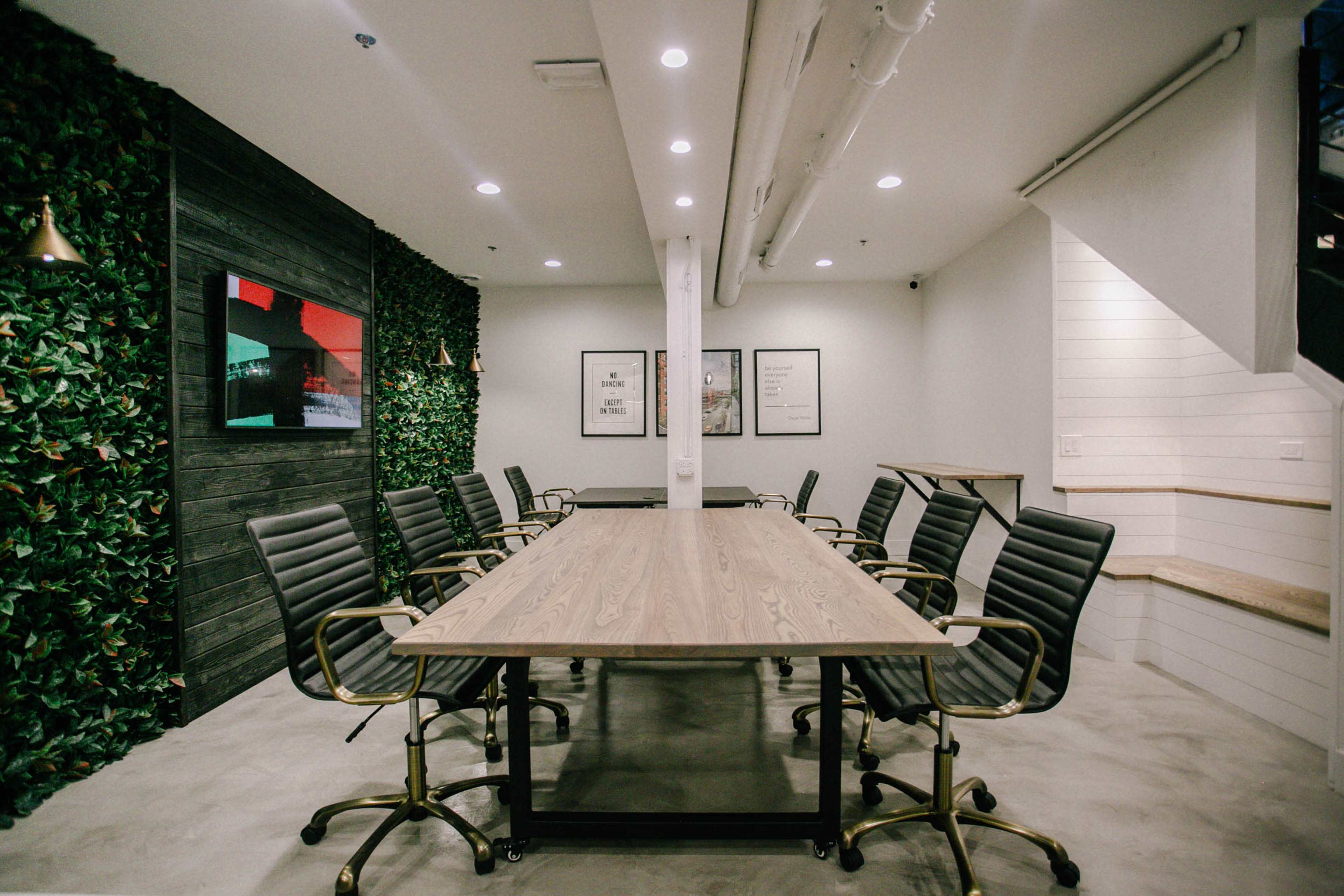 A modern conference room features a large wooden table surrounded by black chairs, with greenery on one wall and framed artwork on another.