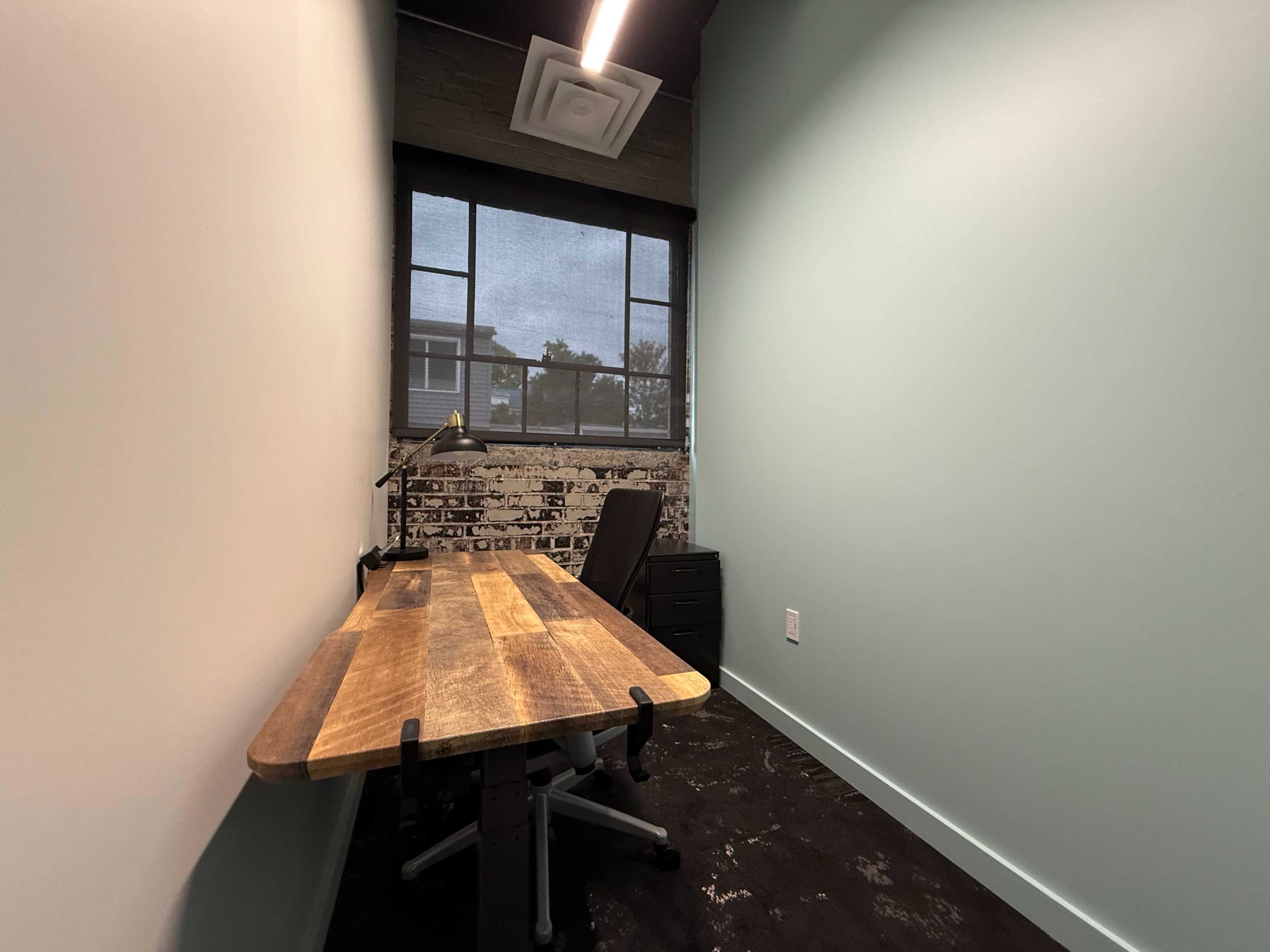 The image shows a small office space with a wooden desk, a black chair, a desk lamp, and a window featuring a view of an outdoor area.
