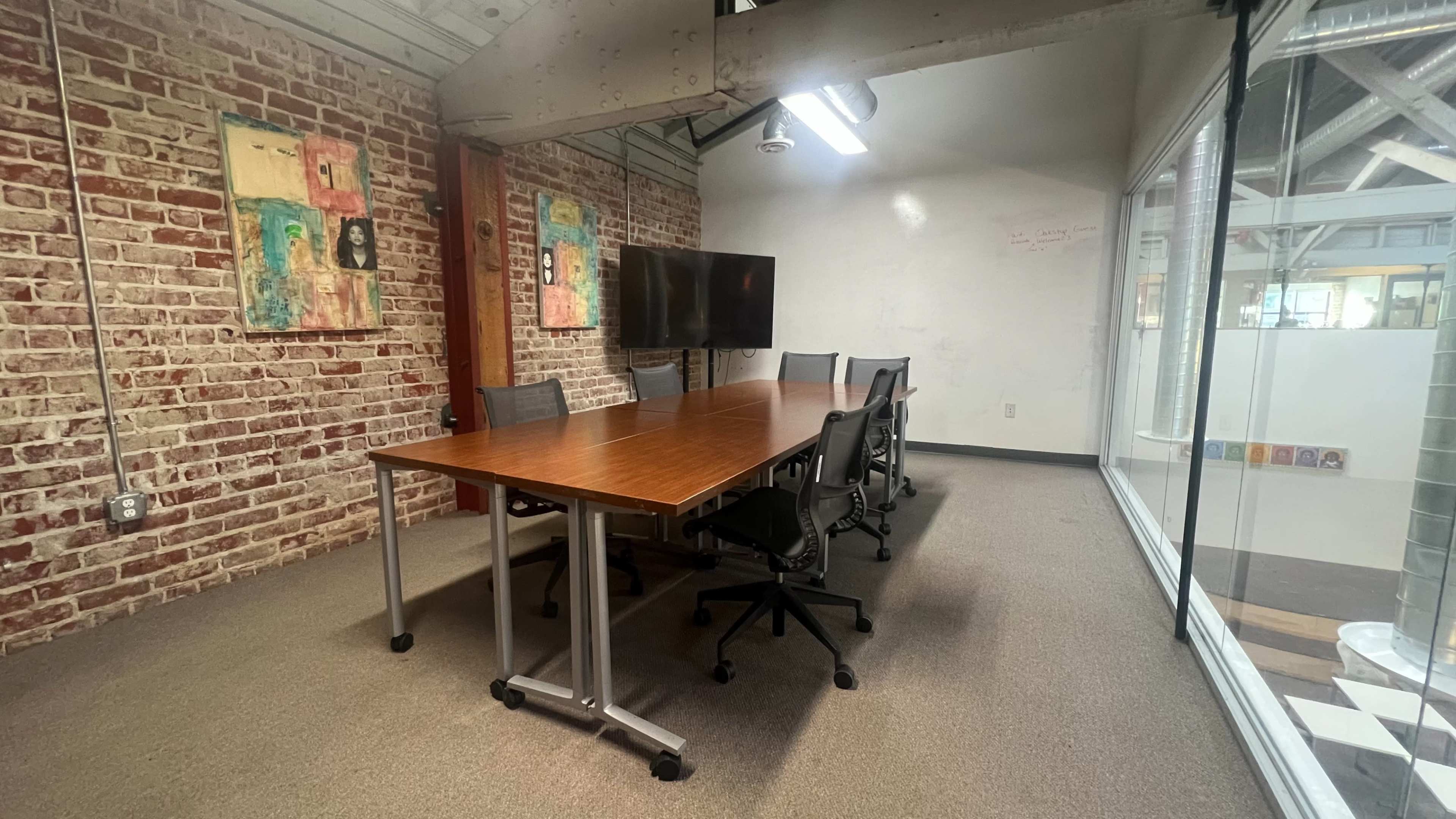 A conference room features a long wooden table surrounded by chairs, with a large screen and exposed brick walls.