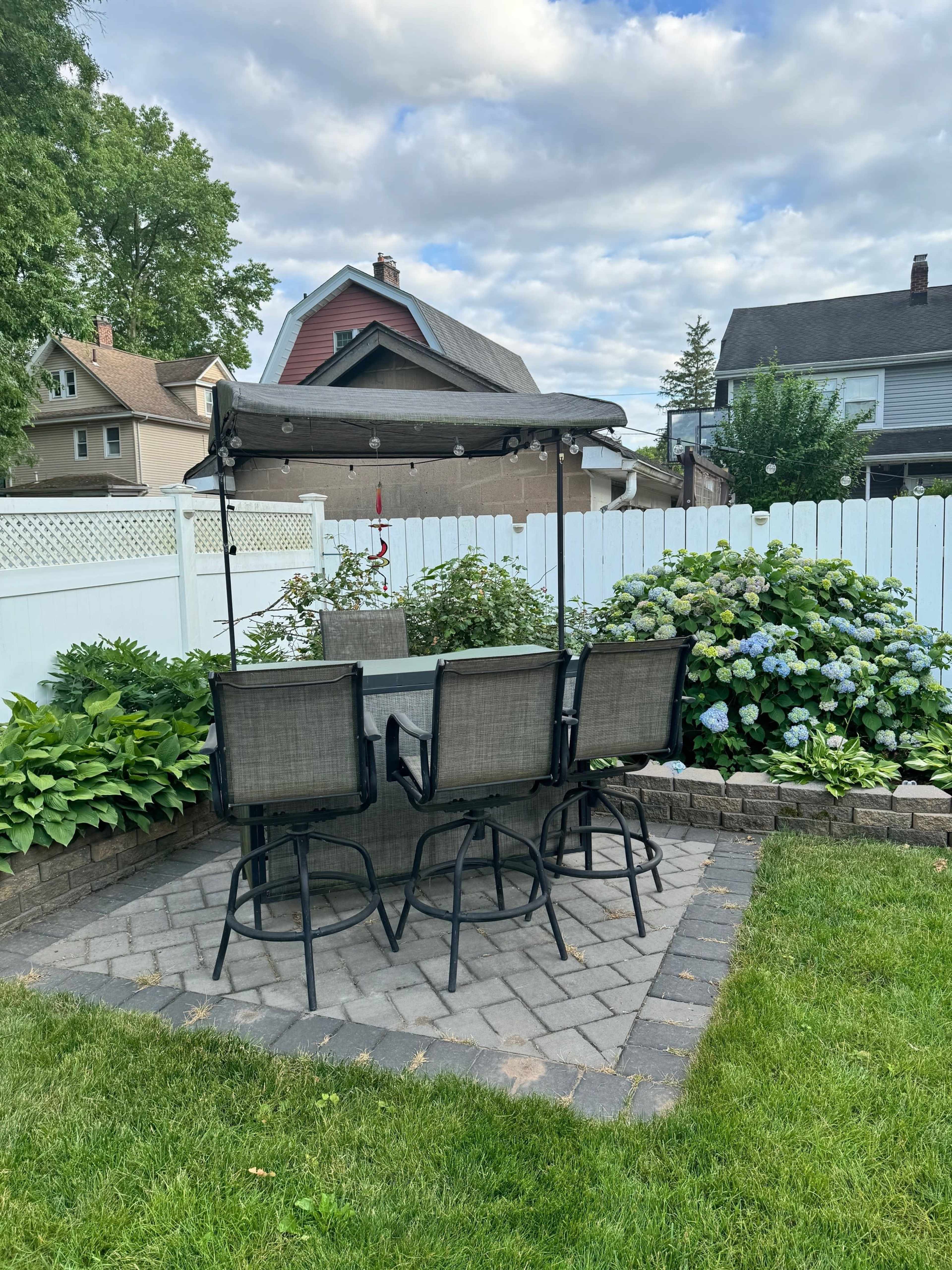 A stone patio with four metal chairs and a table is surrounded by lush greenery and hydrangea bushes, with a white fence and houses visible in the background.