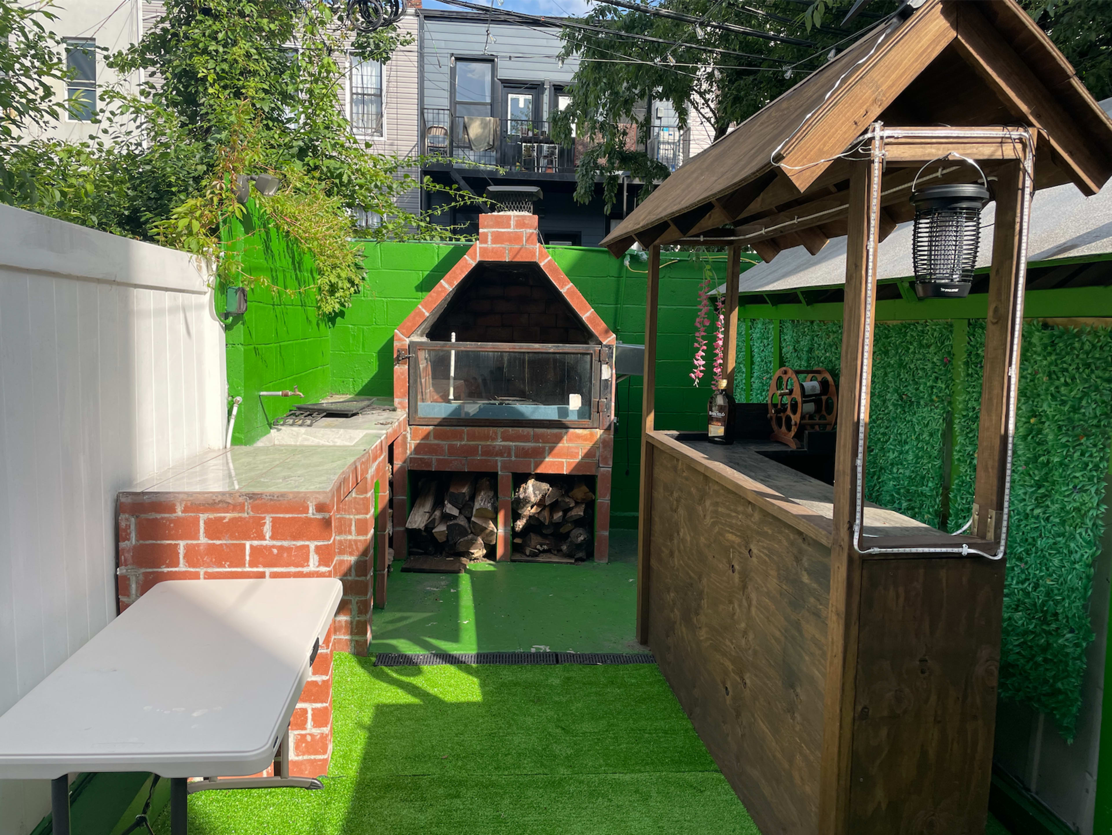 An outdoor space featuring a brick pizza oven, a wooden bar, and a table on artificial grass, surrounded by greenery and fences.