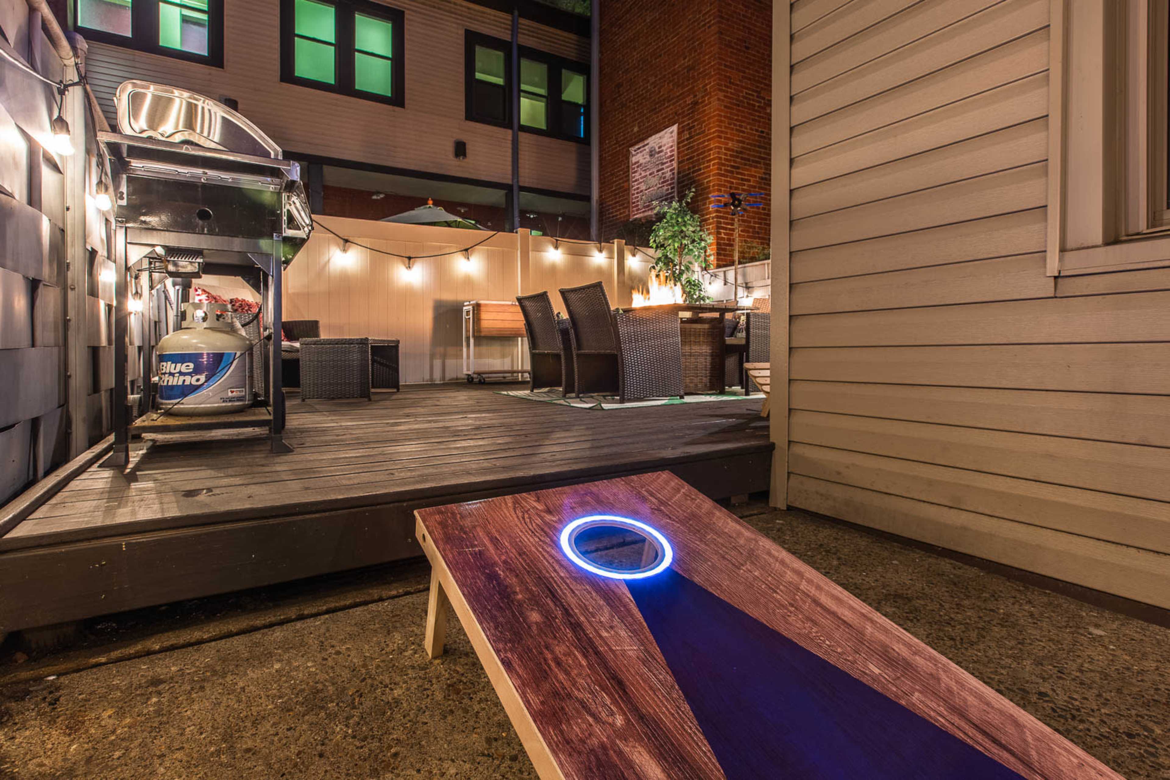 A backyard space with a cornhole game setup, a grill, outdoor dining furniture, and ambient lighting.