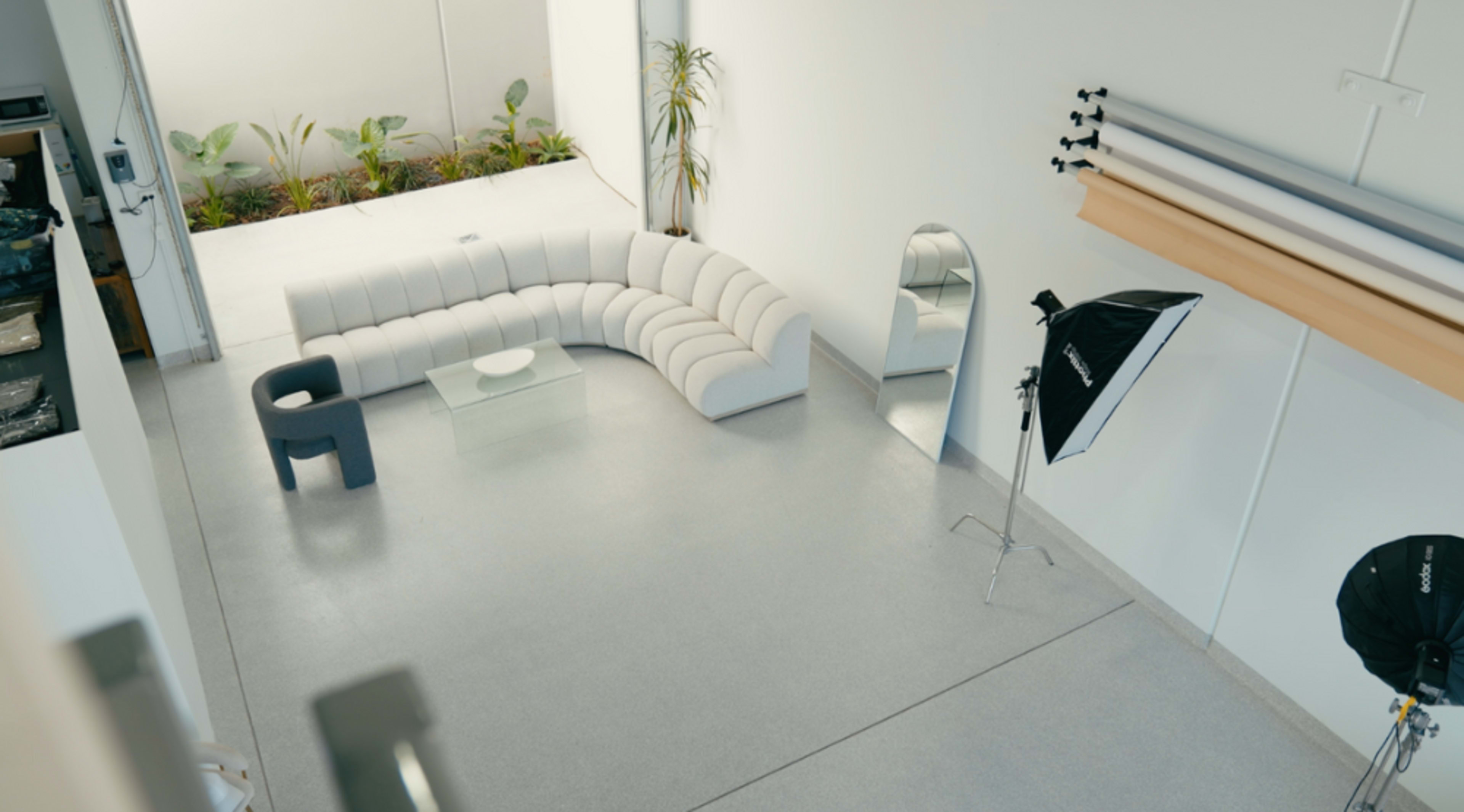 A spacious room with a curved white sofa, a glass coffee table, a large mirror, and a photography setup including a softbox light and backdrops.