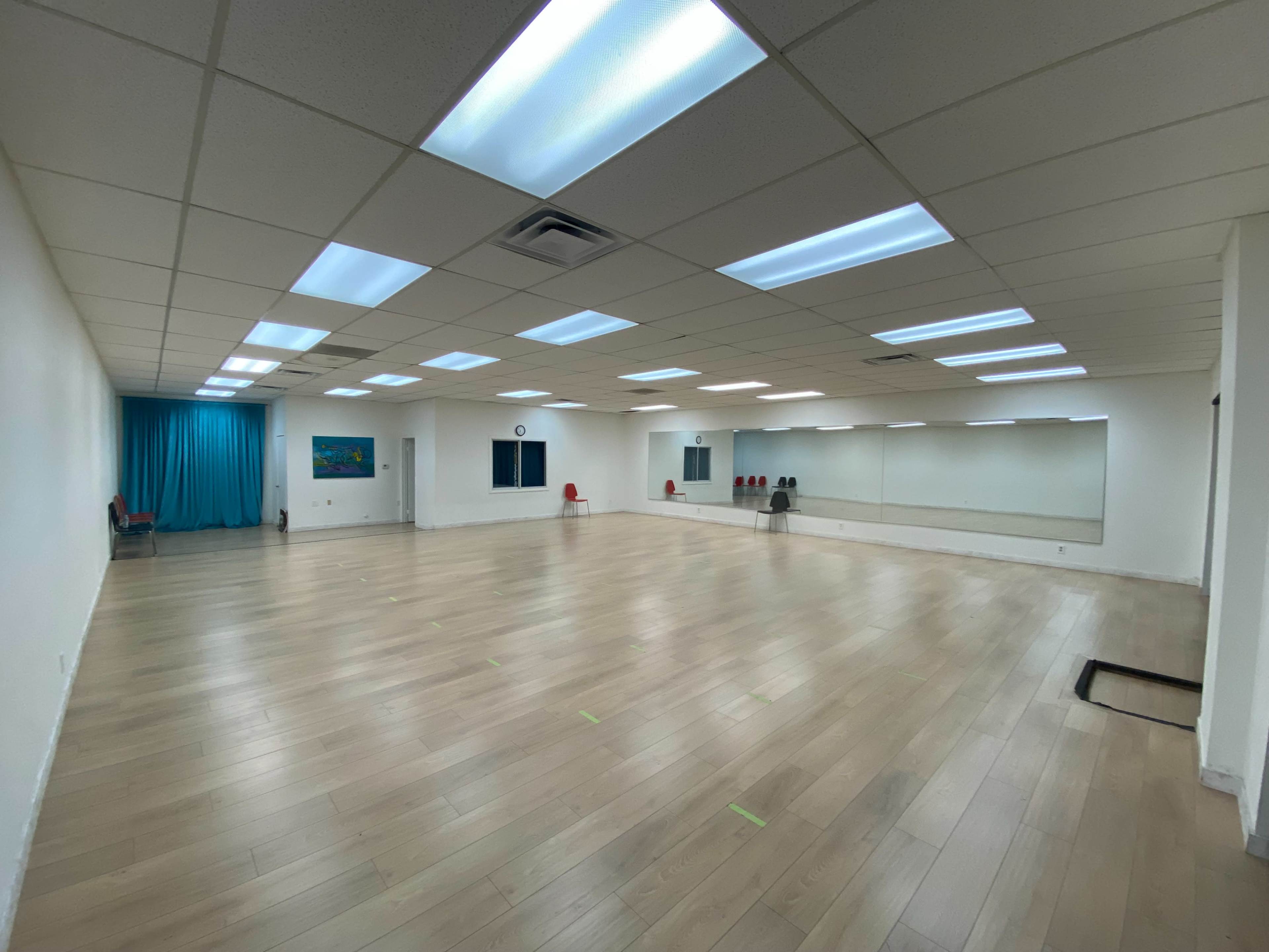 A spacious, well-lit room with a wooden floor, mirrors on one wall, and a blue curtain at the far end.