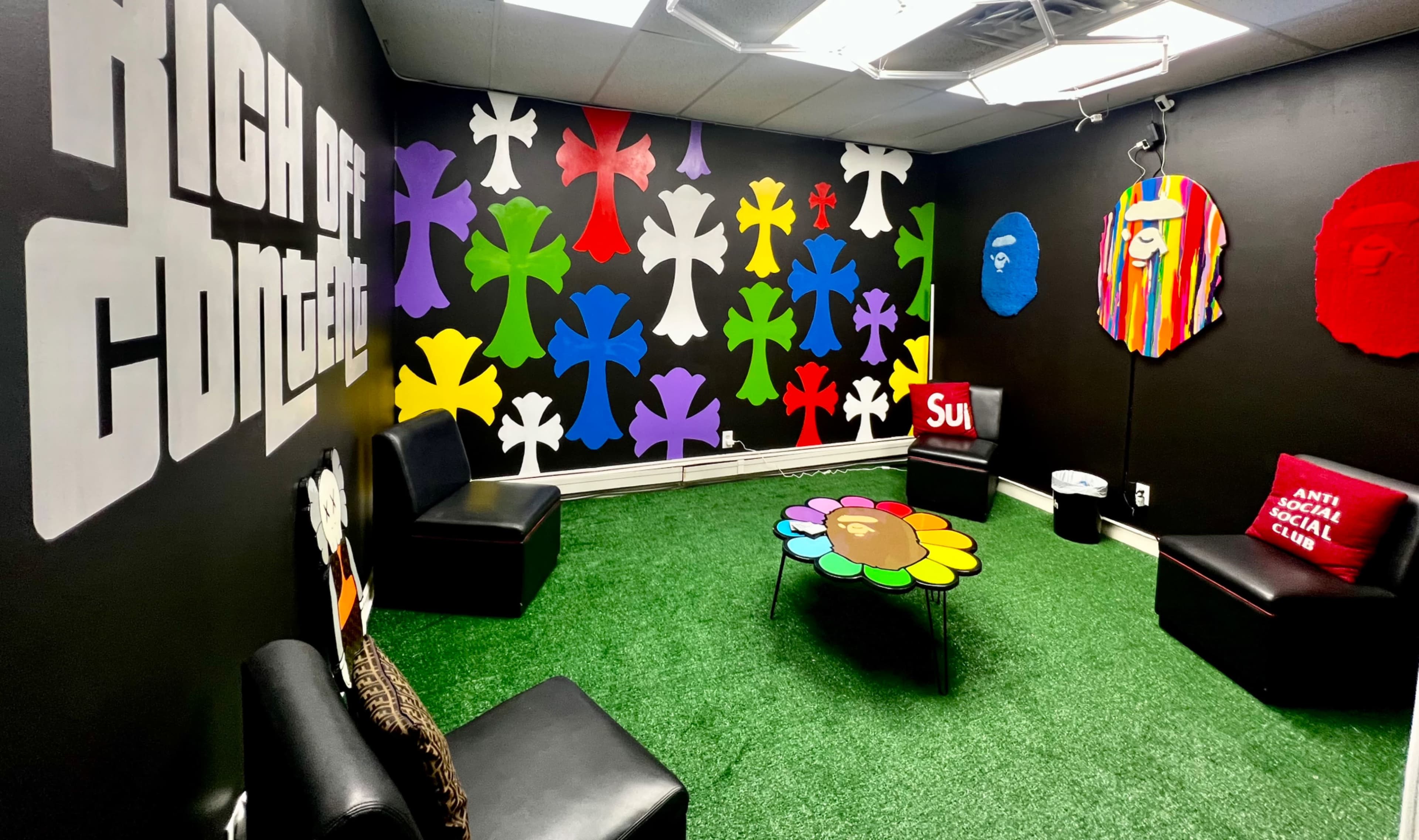 The image shows a vibrant, creative lounge area with black walls adorned with colorful flower-shaped decorations and a flower-patterned coffee table on artificial grass flooring.