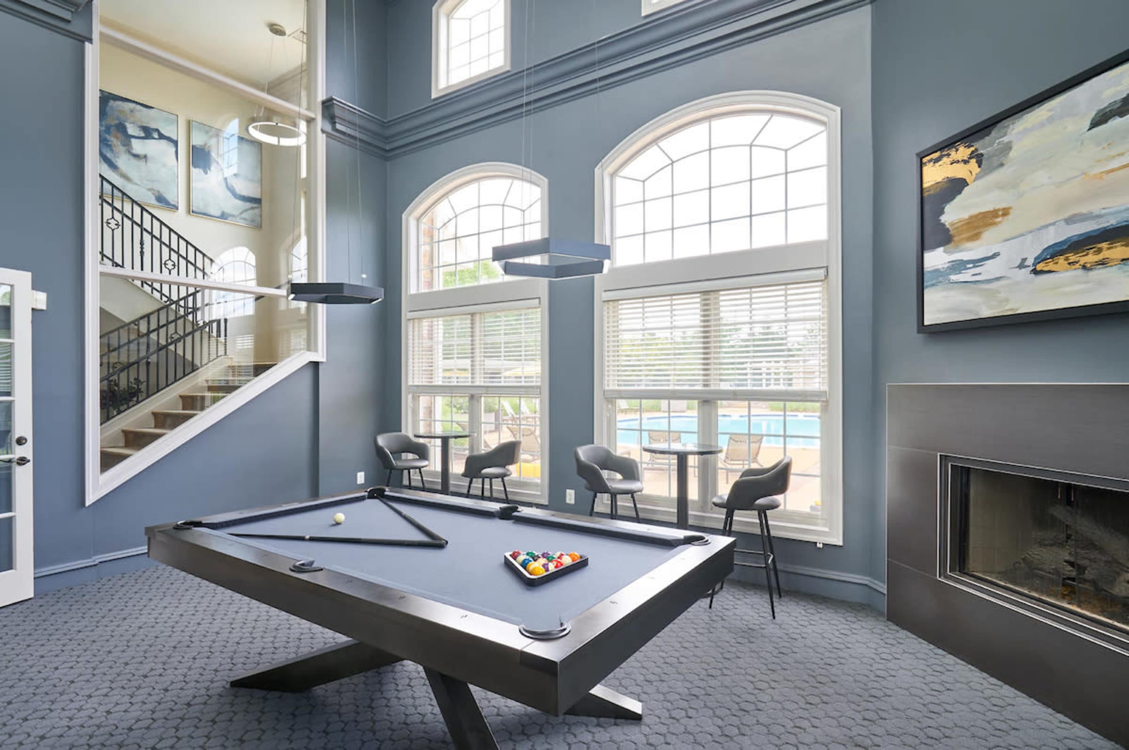 A modern room with a pool table, large windows overlooking a pool area, a staircase, and contemporary seating arrangements.