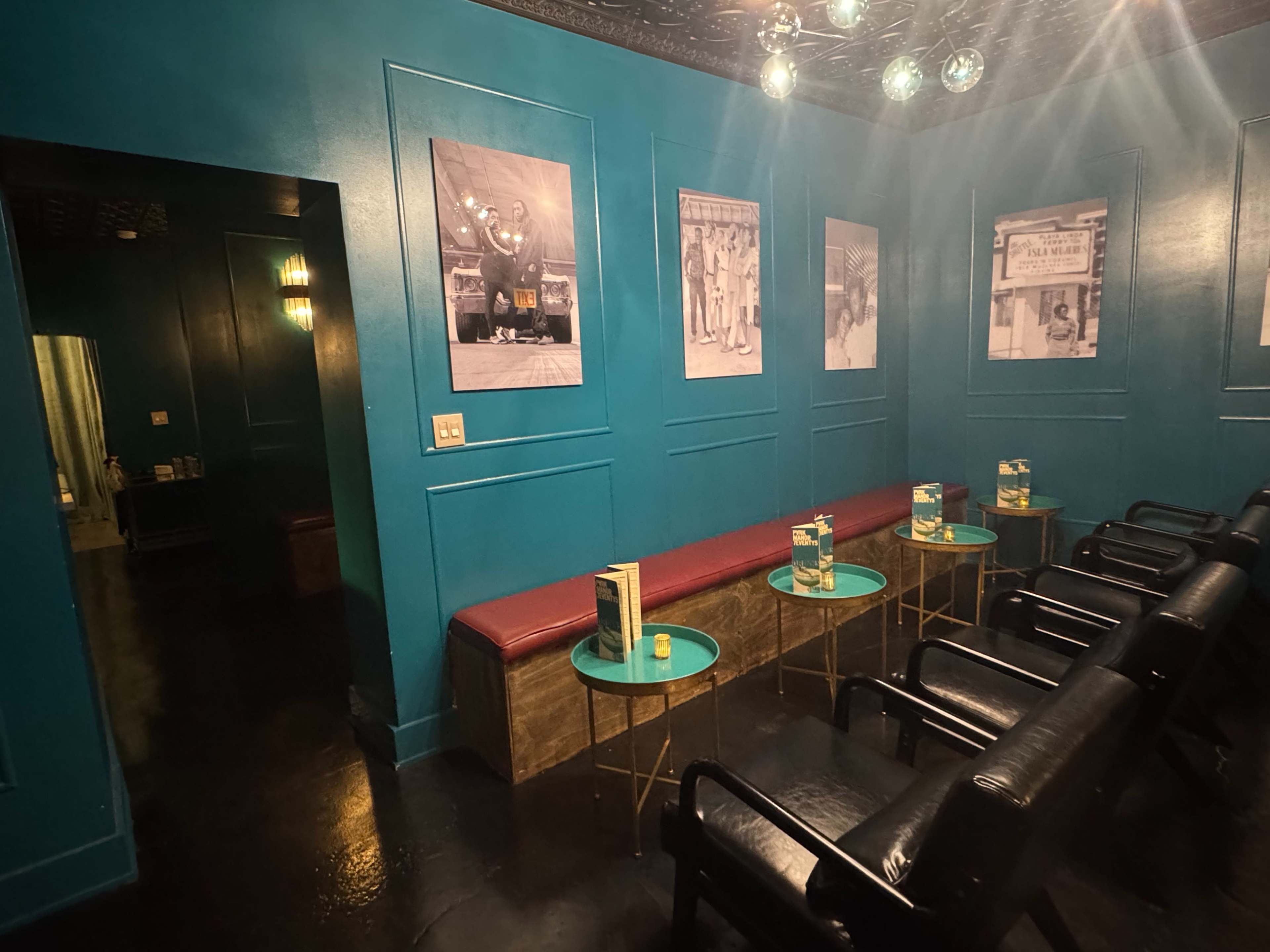 The image shows a waiting area featuring teal walls adorned with black-and-white photographs, with a red bench and small glass-top tables accompanied by black chairs.