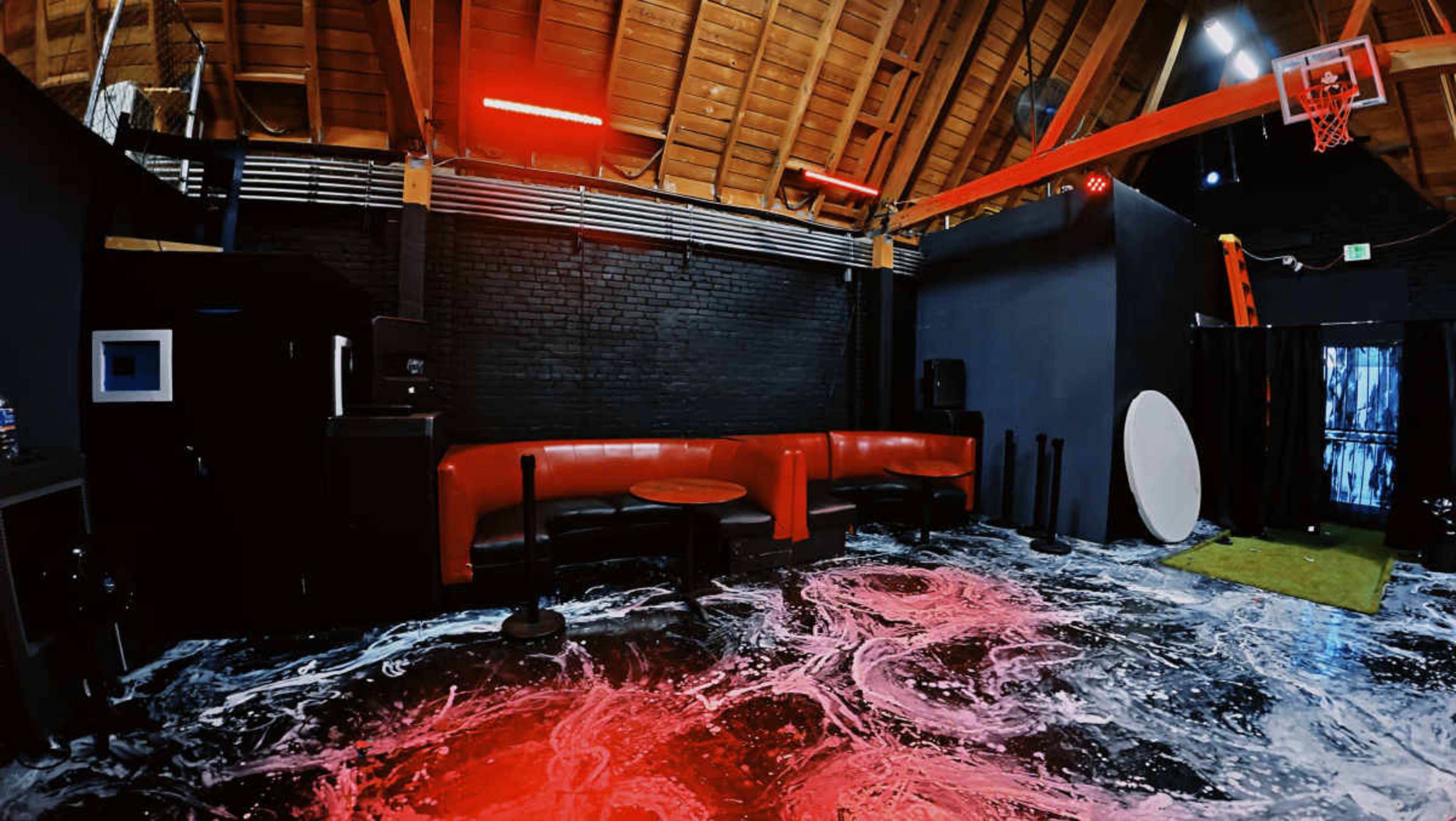 The image shows a spacious room with a wooden ceiling, featuring a black wall, red couches, and a colorful floor design, along with a basketball hoop in the background.