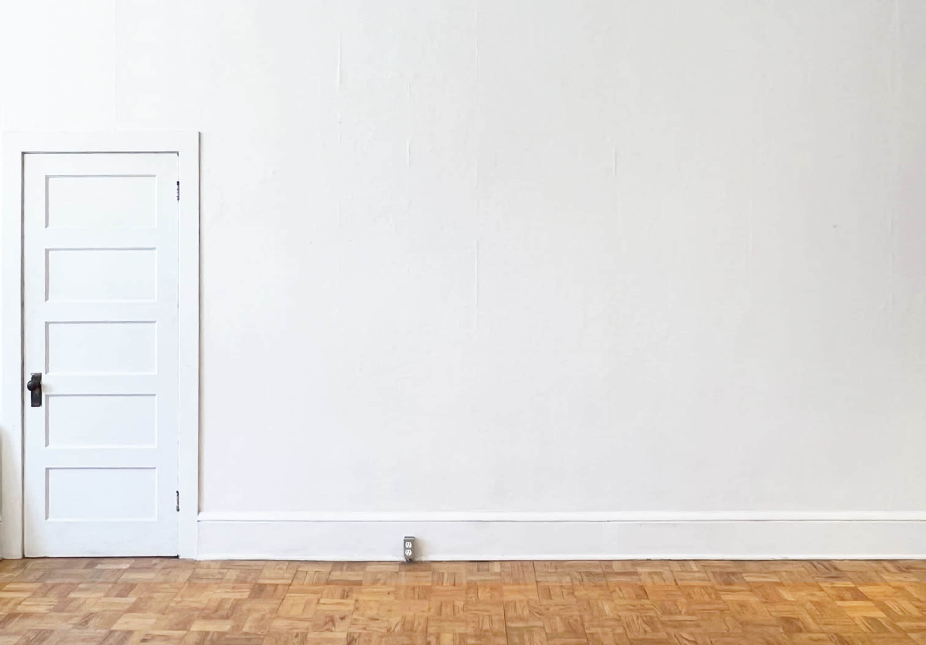 A plain white wall features a closed, white door with five panels and a wooden floor made of parquet.