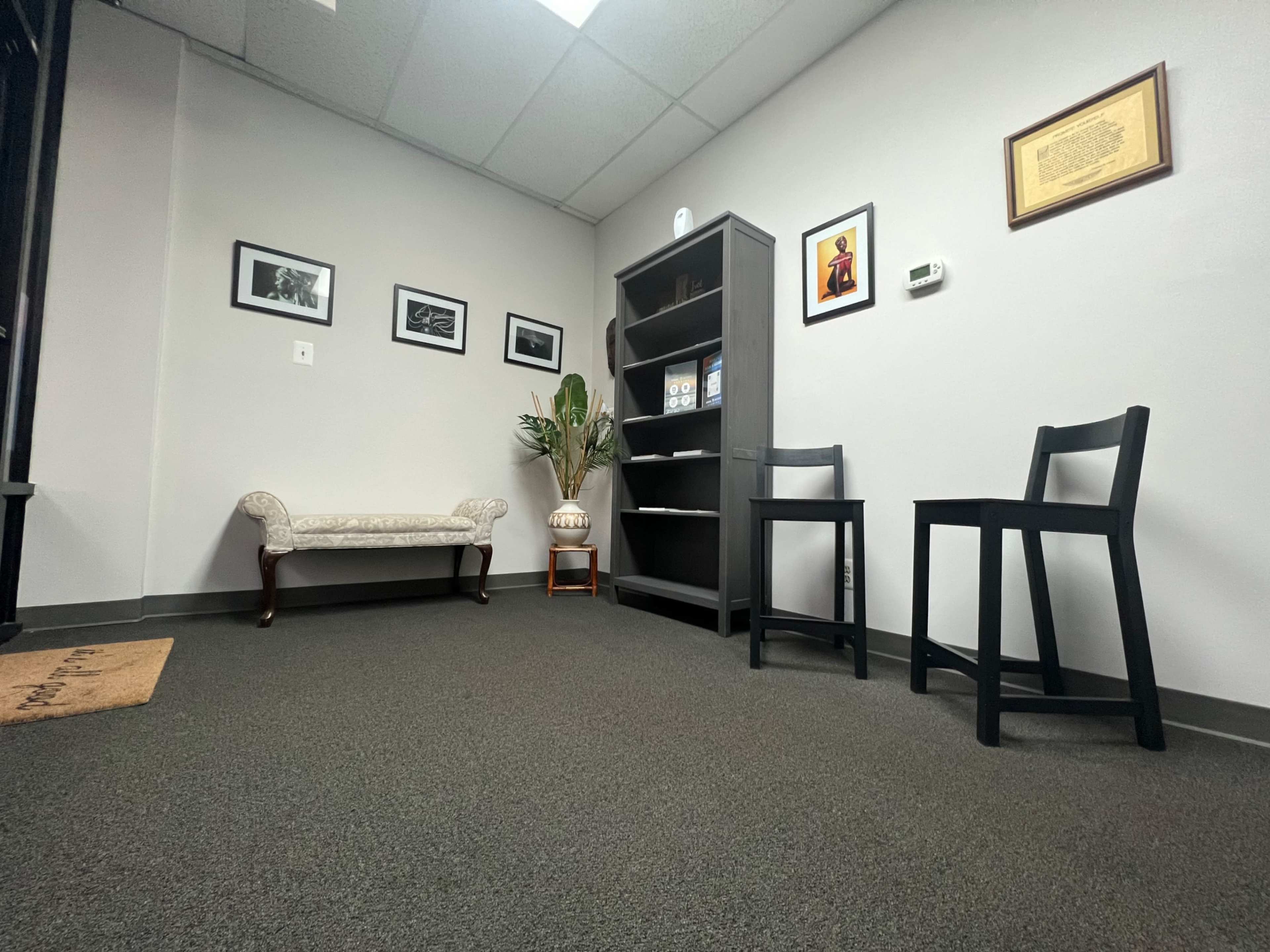 A small, minimalist office space with a decorative chair, a bookshelf, and framed pictures on the walls.