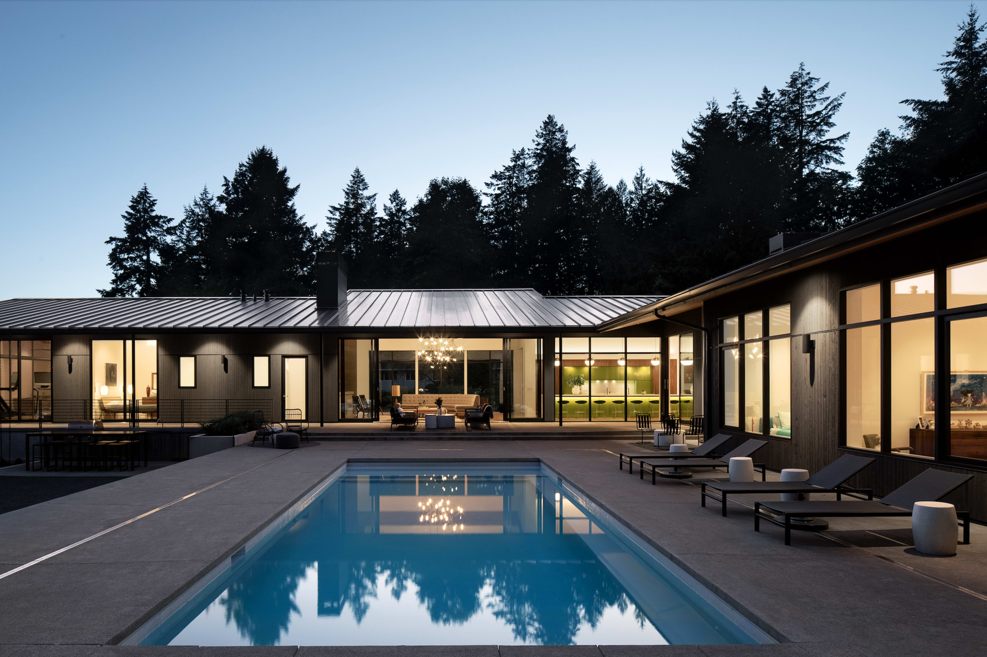 A modern house with large windows surrounds a crystal-clear swimming pool, set against a backdrop of tall trees at twilight.