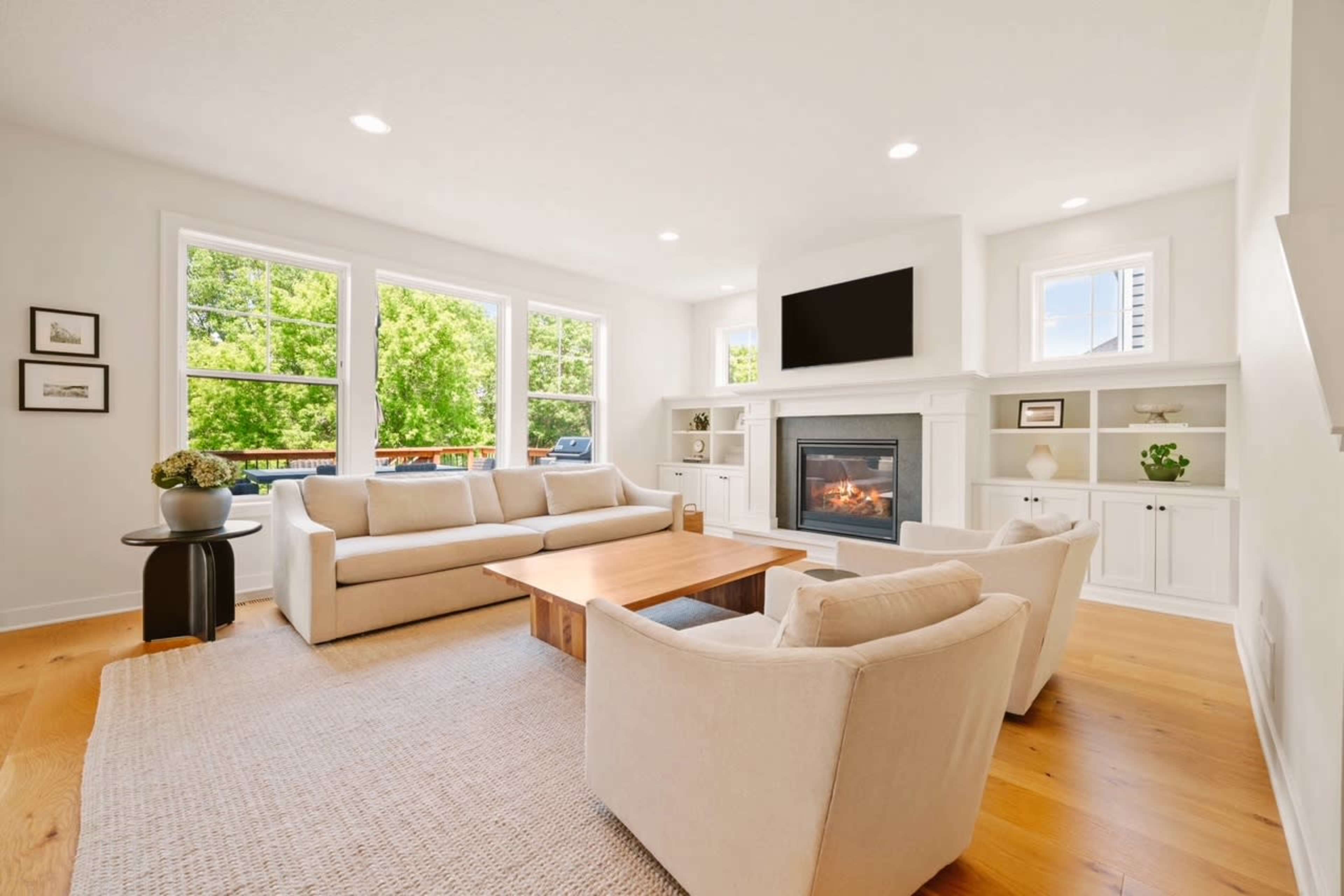 The living room features a beige sofa set arranged around a wooden coffee table, with large windows allowing natural light and a fireplace integrated into the wall.