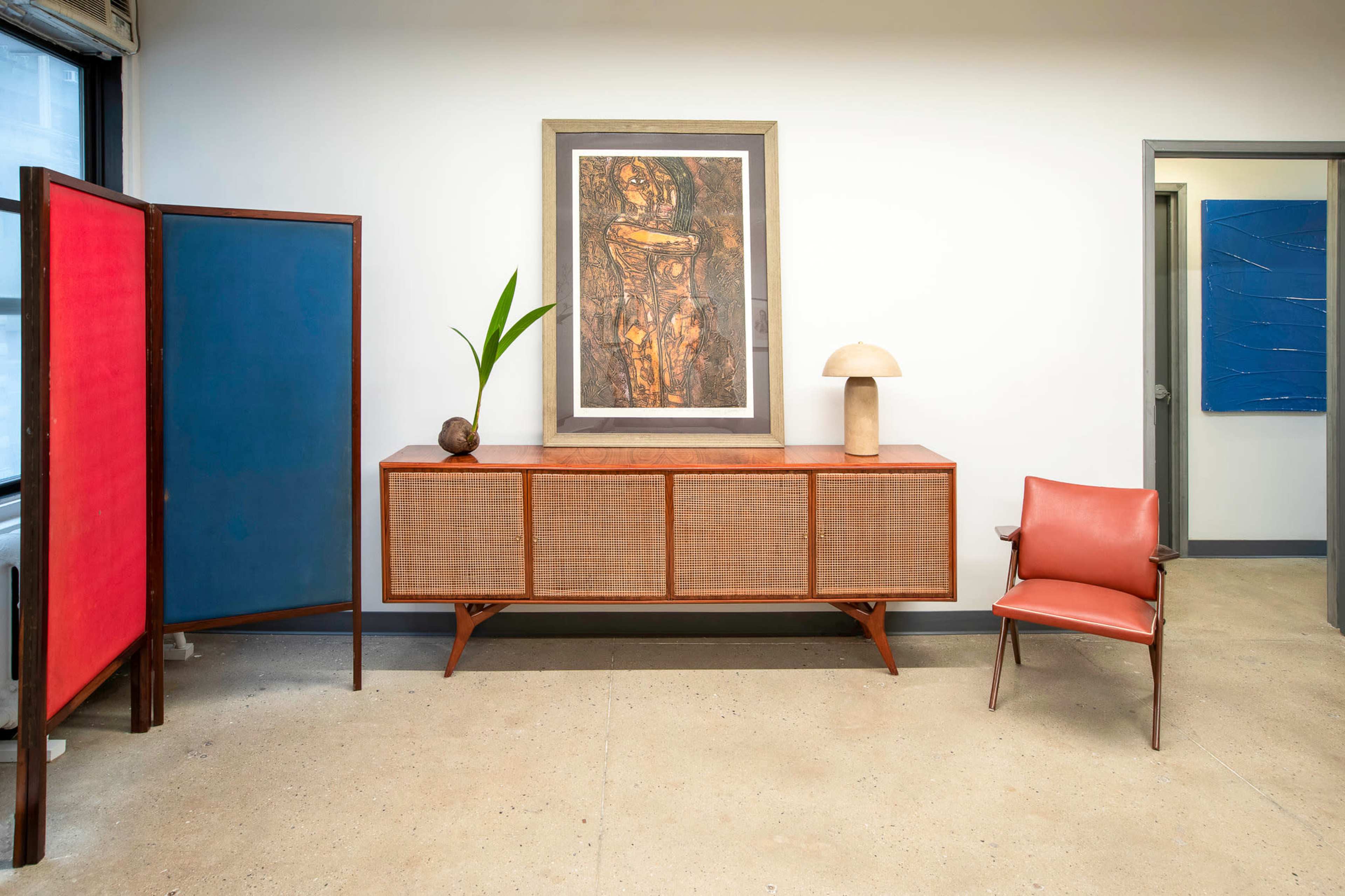 The interior scene features a wooden sideboard with a decorative lamp and plant, flanked by a red and blue room divider, alongside a leather chair and a piece of artwork on the wall.