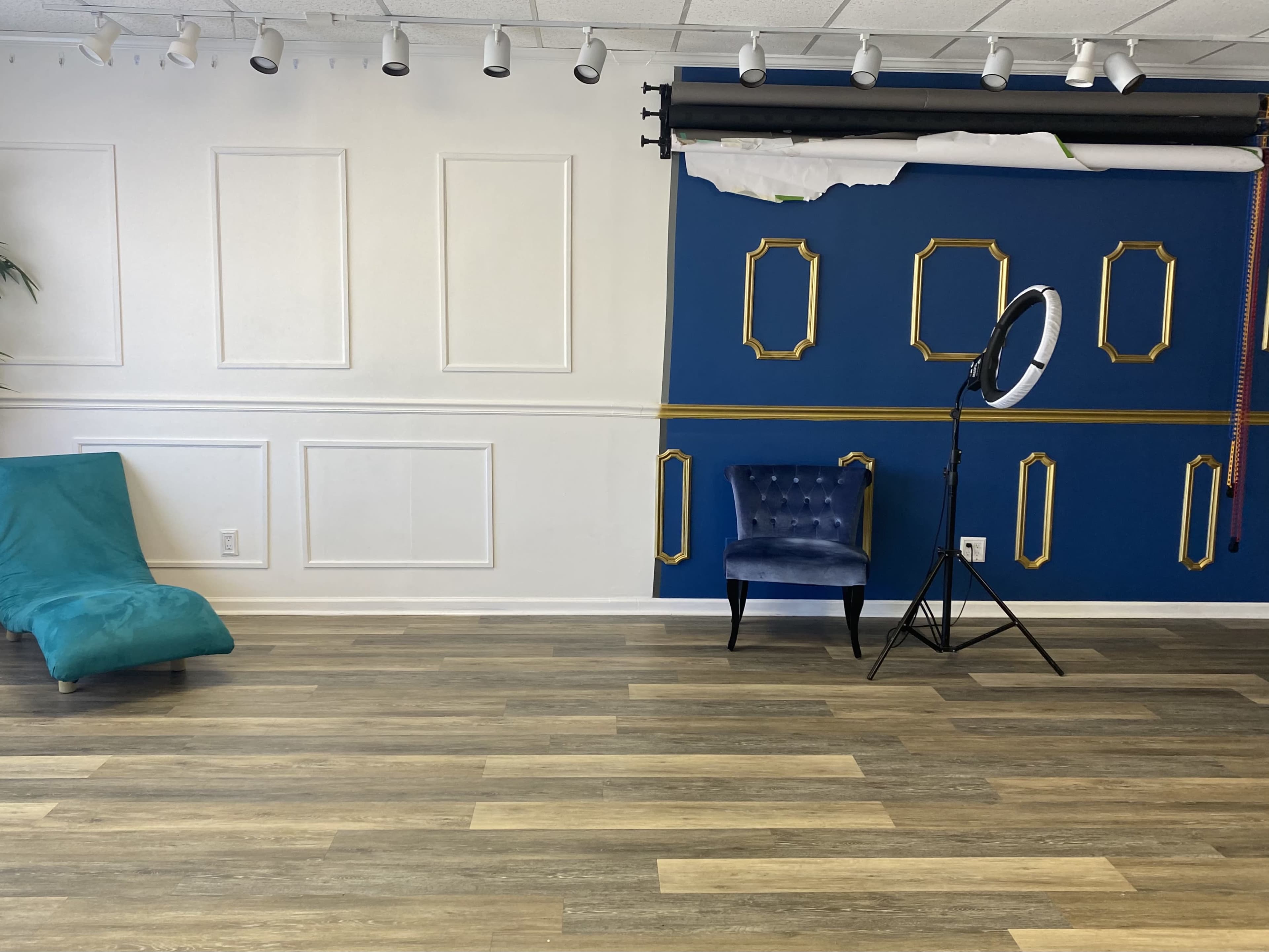 A spacious room with a blue accent wall featuring decorative frames, a turquoise chair, a blue upholstered chair, and a lighting setup on a tripod.