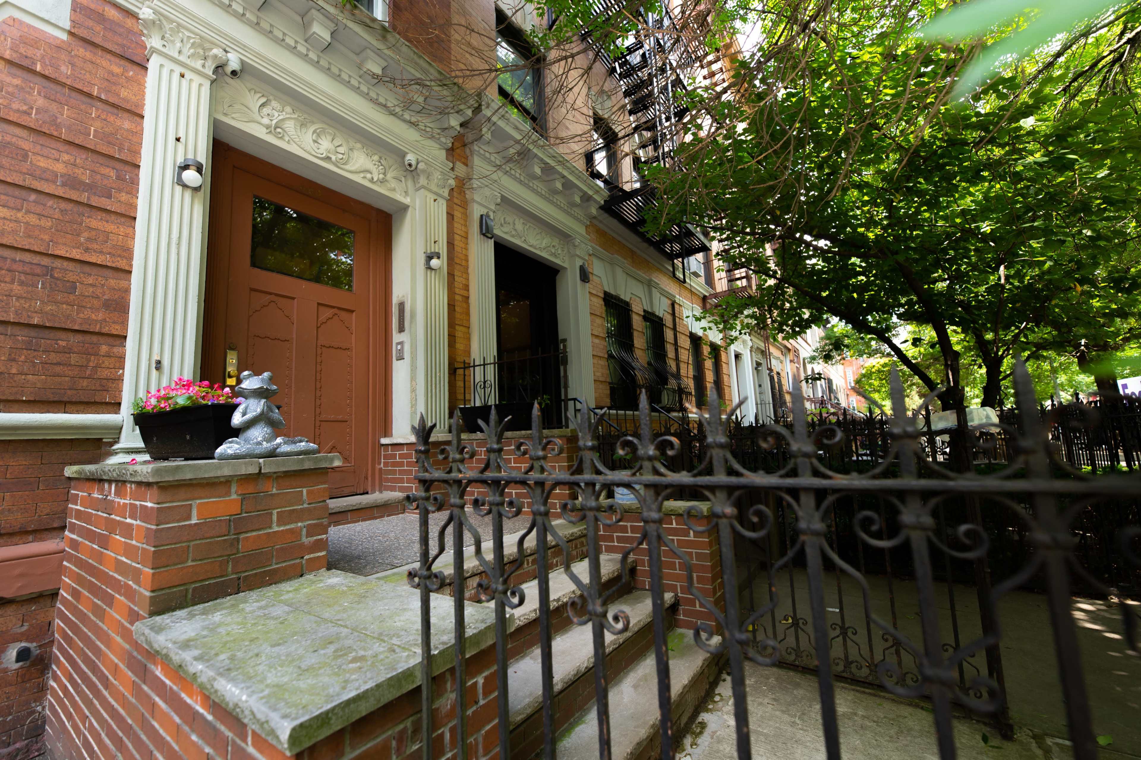A brownstone building with decorative features, a set of stairs leading to an entrance, and a wrought-iron fence surrounded by greenery.