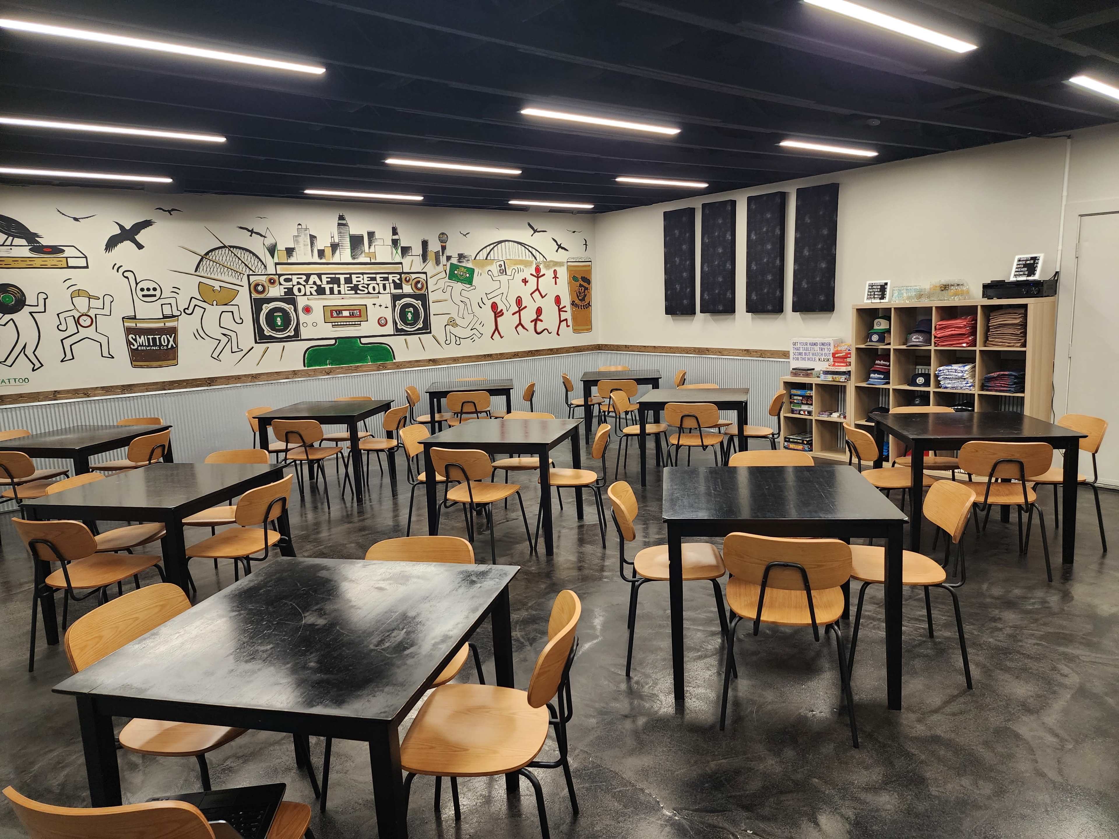 The image shows a spacious café with black tables and wooden chairs arranged neatly, decorated with a mural on the wall and shelves filled with colorful items.
