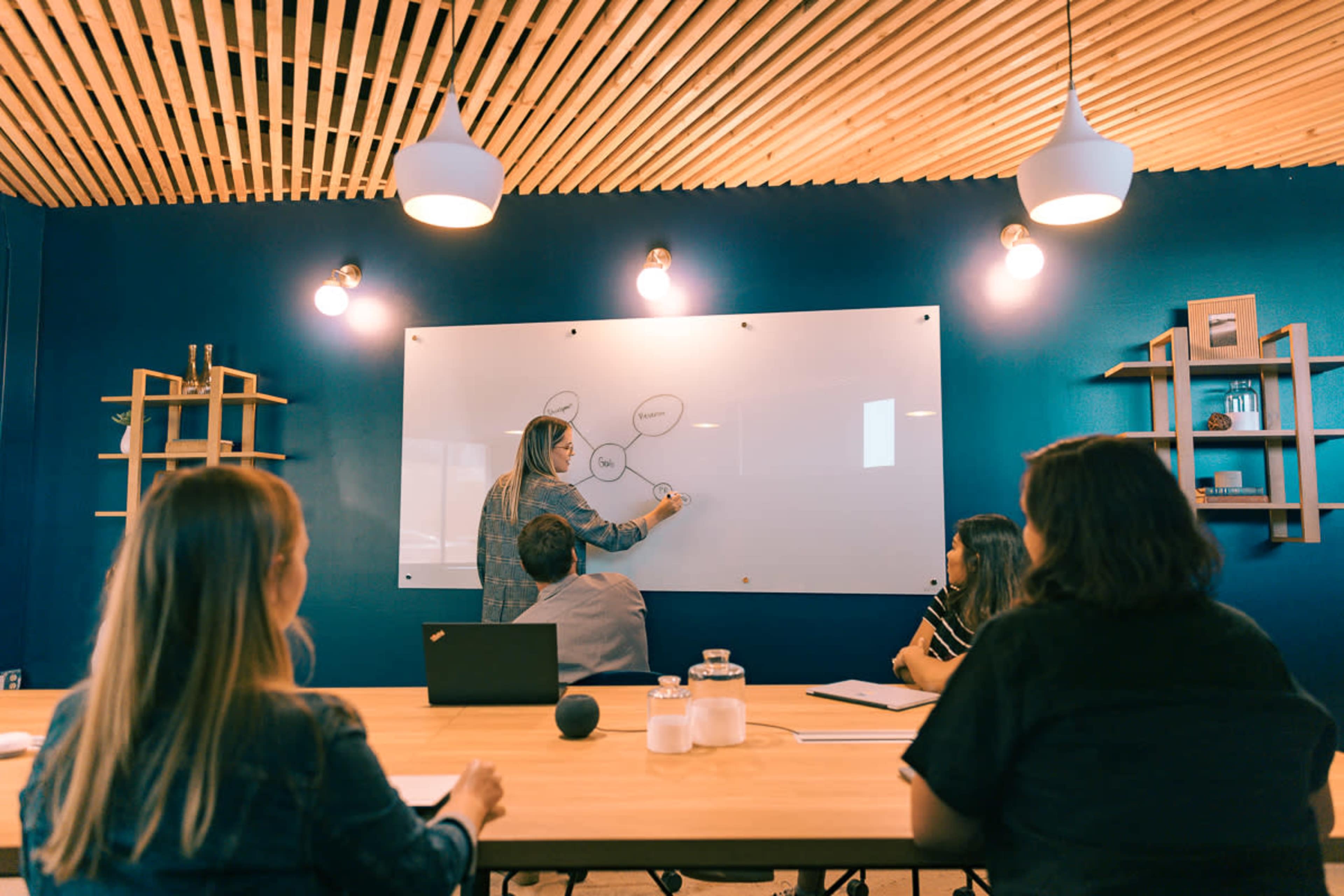 A group of people is seated at a conference table watching a person draw on a whiteboard in a modern meeting room.