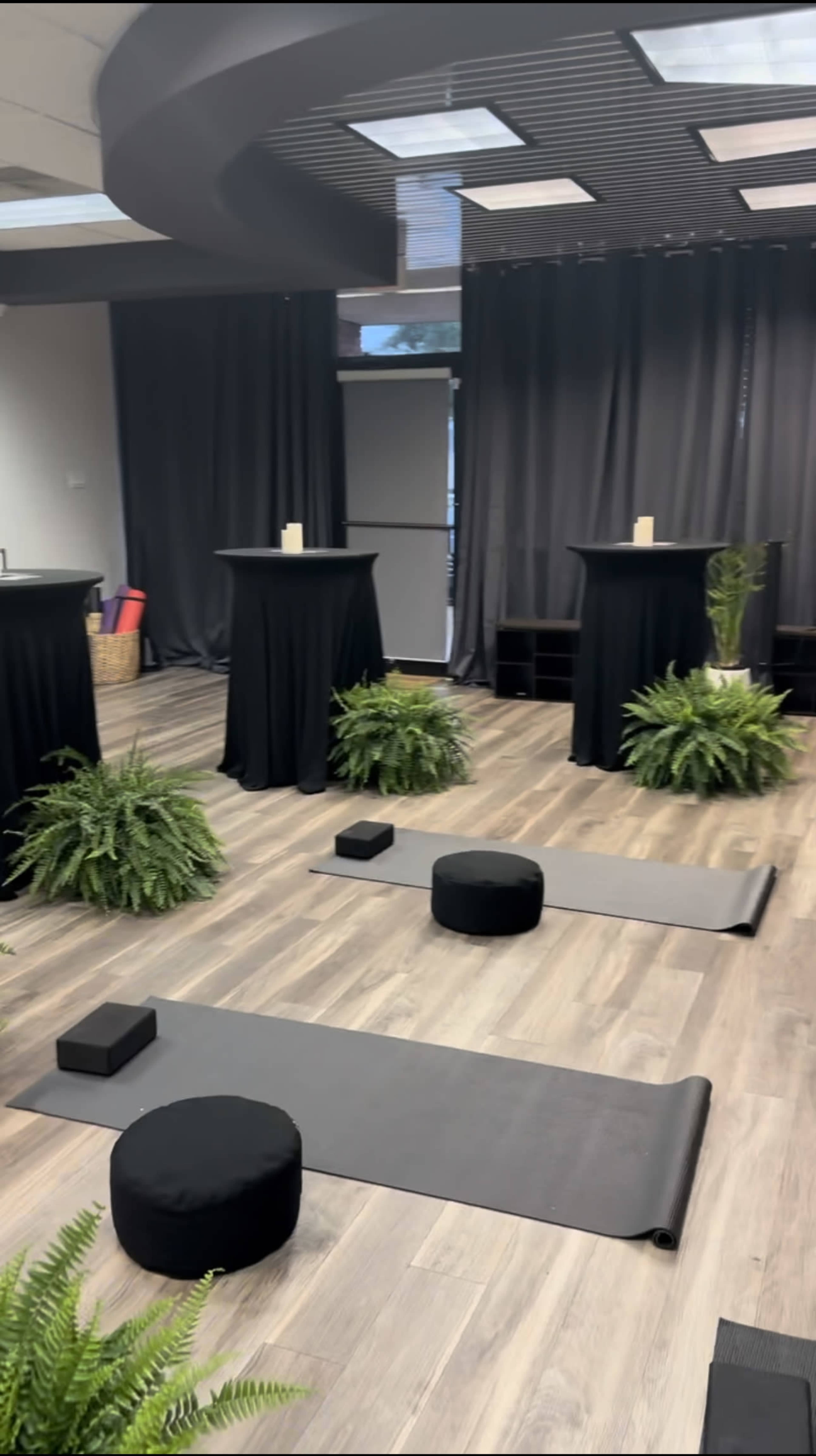 The image depicts a room set up for a yoga or meditation session, featuring black mats, round cushions, pedestal tables, and potted ferns.