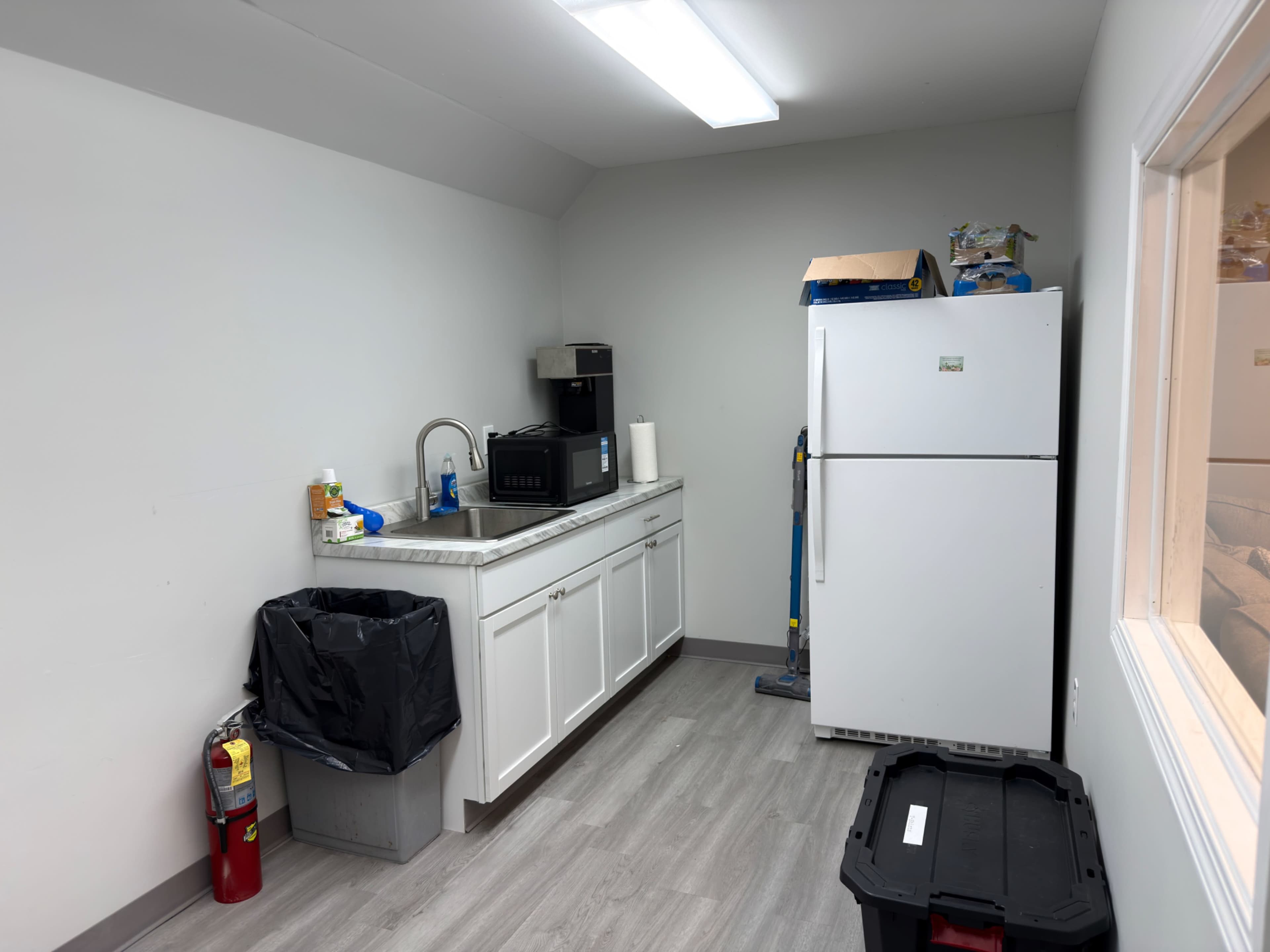 The image shows a small kitchen area with a sink, microwave, and refrigerator, along with a trash can and fire extinguisher.