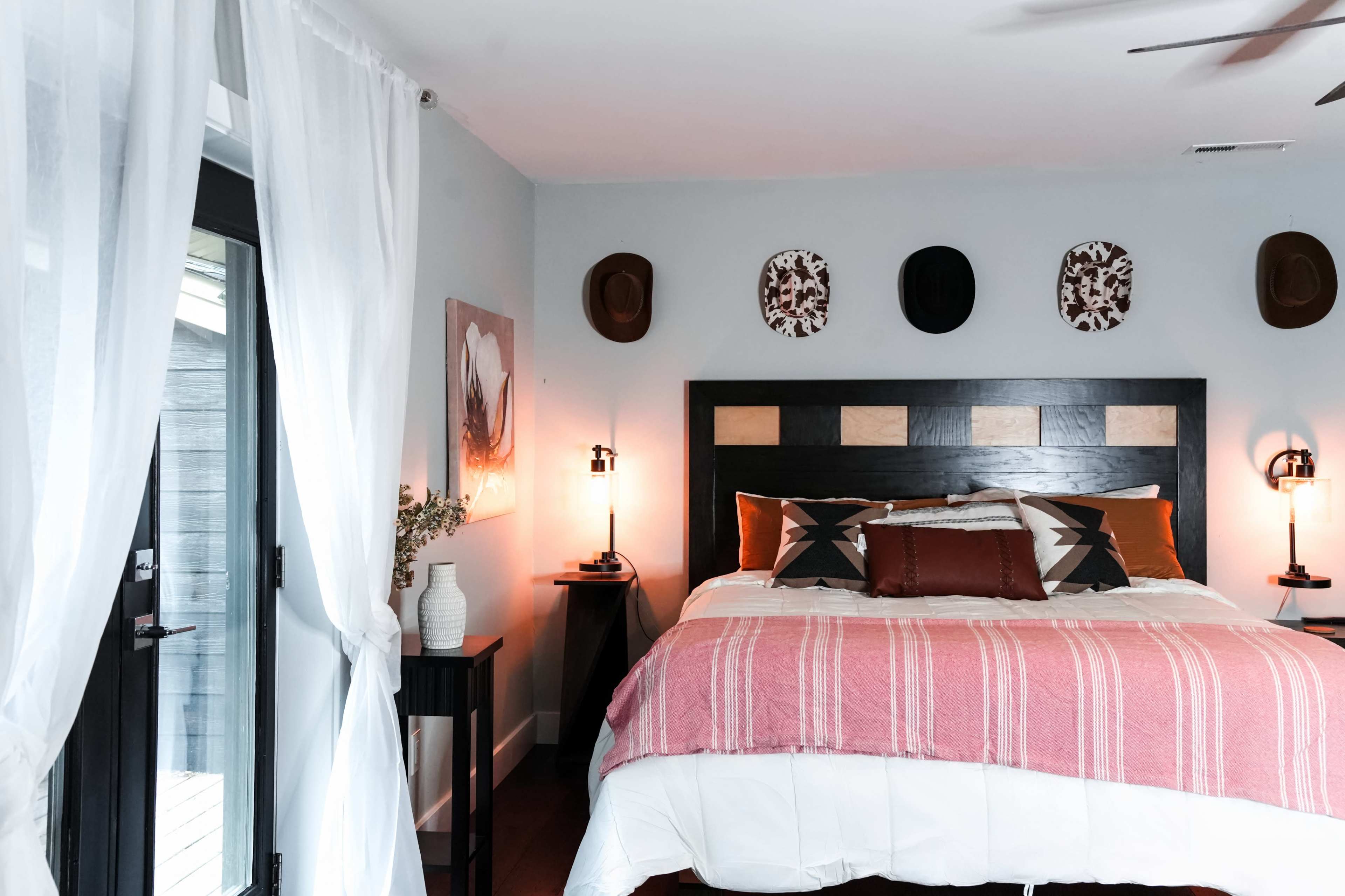 A bedroom features a large bed with patterned pillows and blankets, flanked by two bedside lamps, and adorned with wall decorations and hats above the headboard.