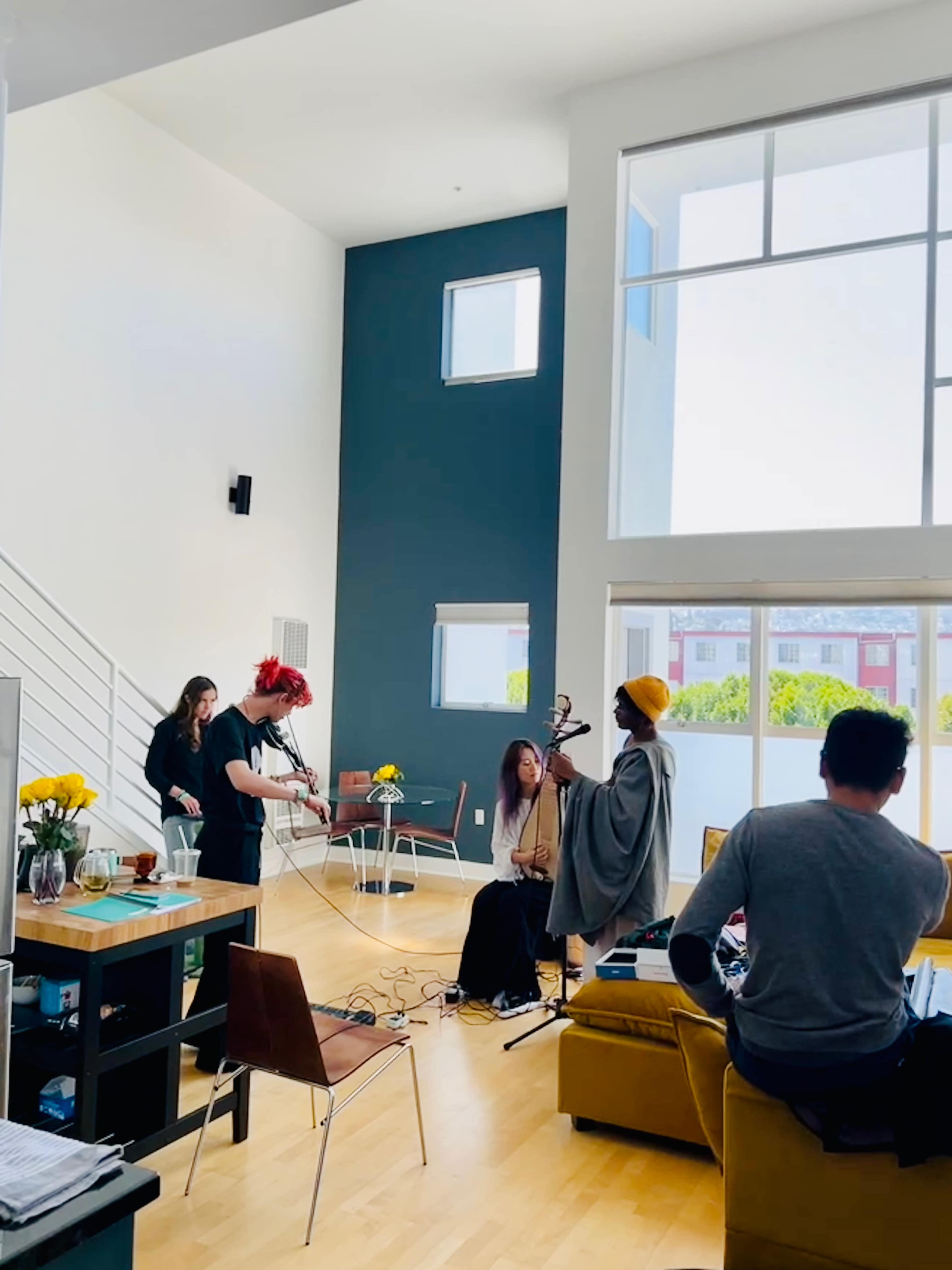 A group of musicians performs in a brightly lit, modern living room with large windows.