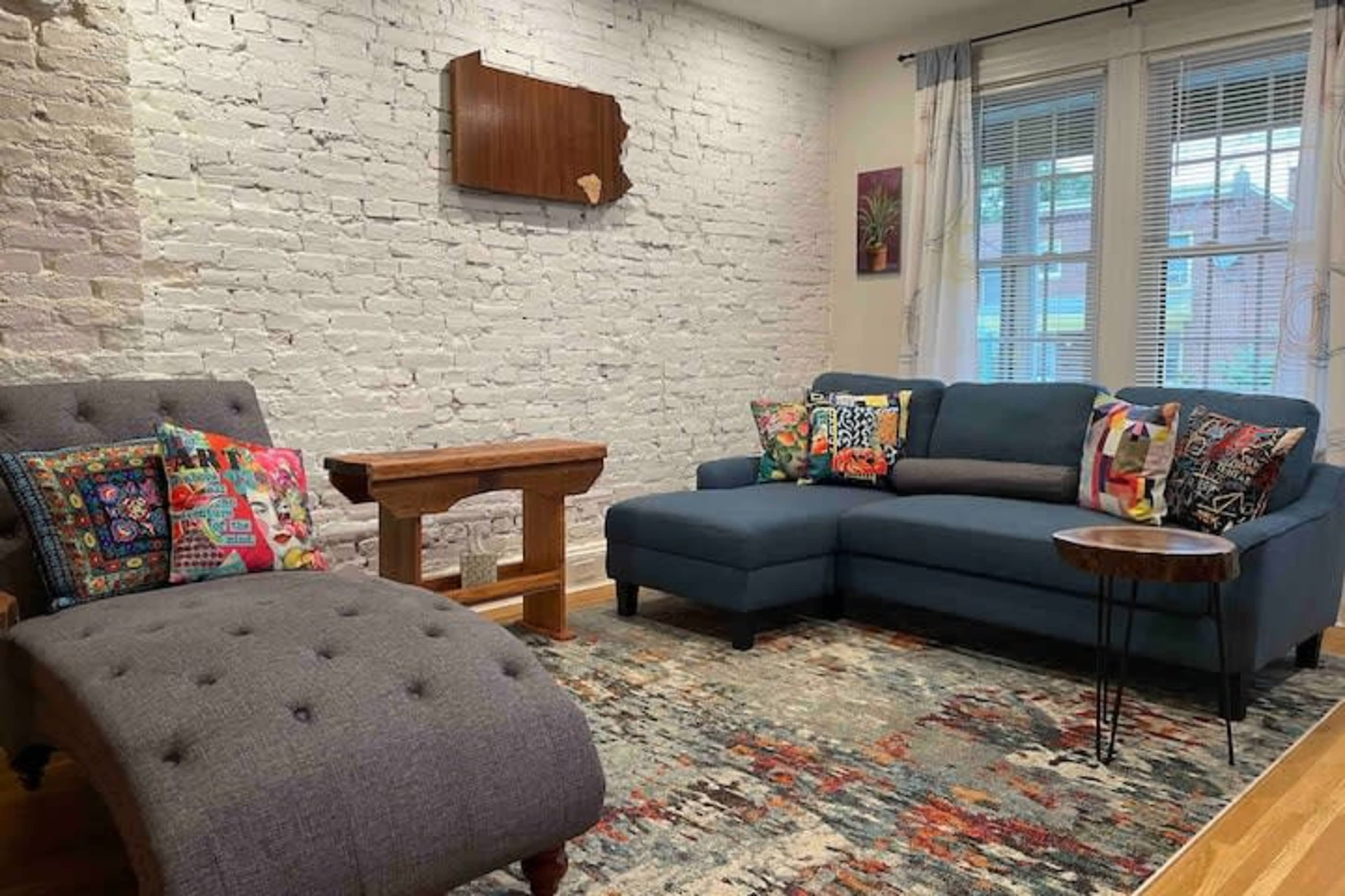 The living room features a blue sectional sofa with colorful cushions, a wooden side table, a gray chaise lounge, and a textured area rug on a hardwood floor against a white brick wall.