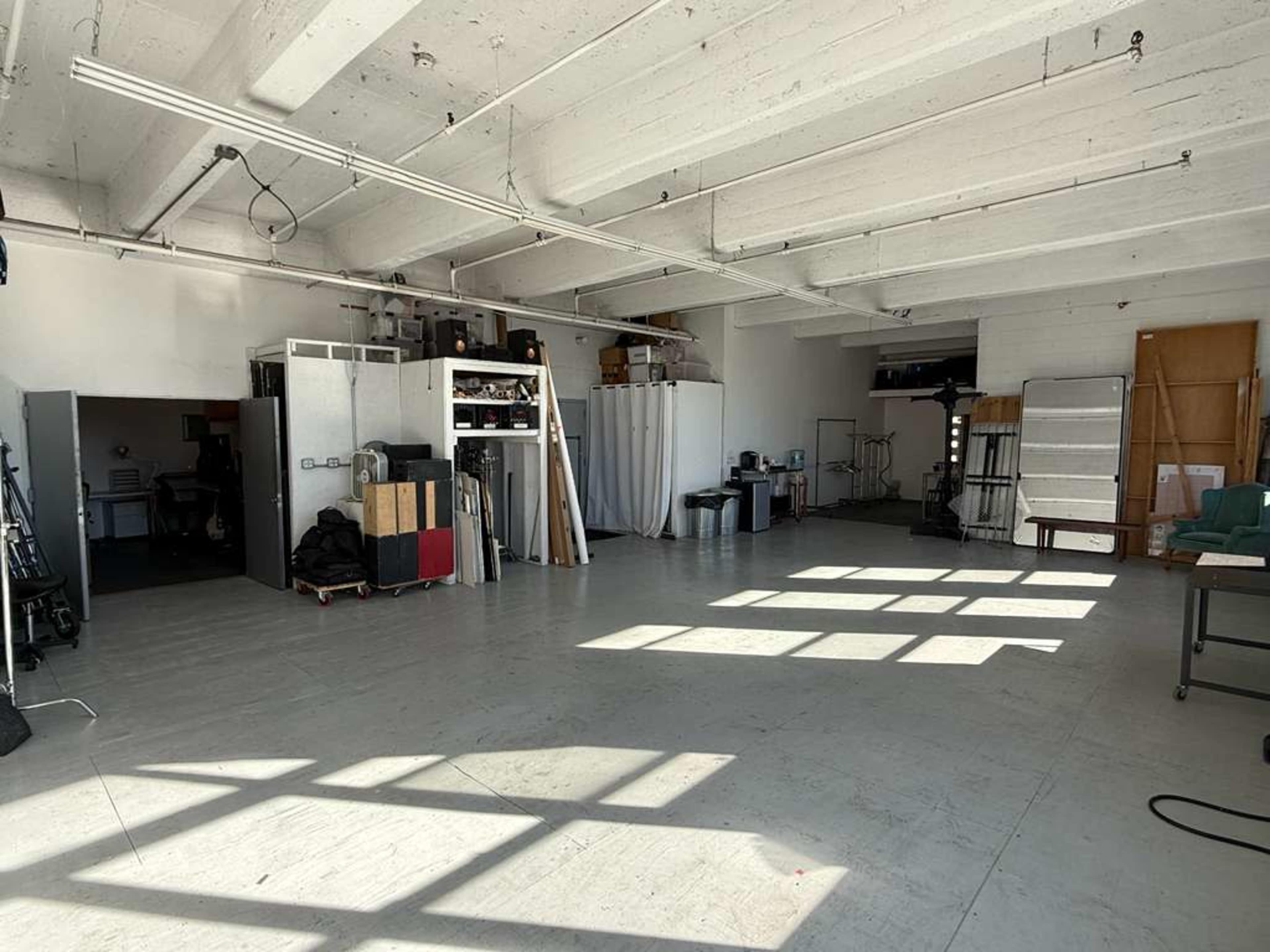 The image shows a spacious studio or workshop area with concrete flooring, high ceilings, and various equipment and materials stored along the walls.