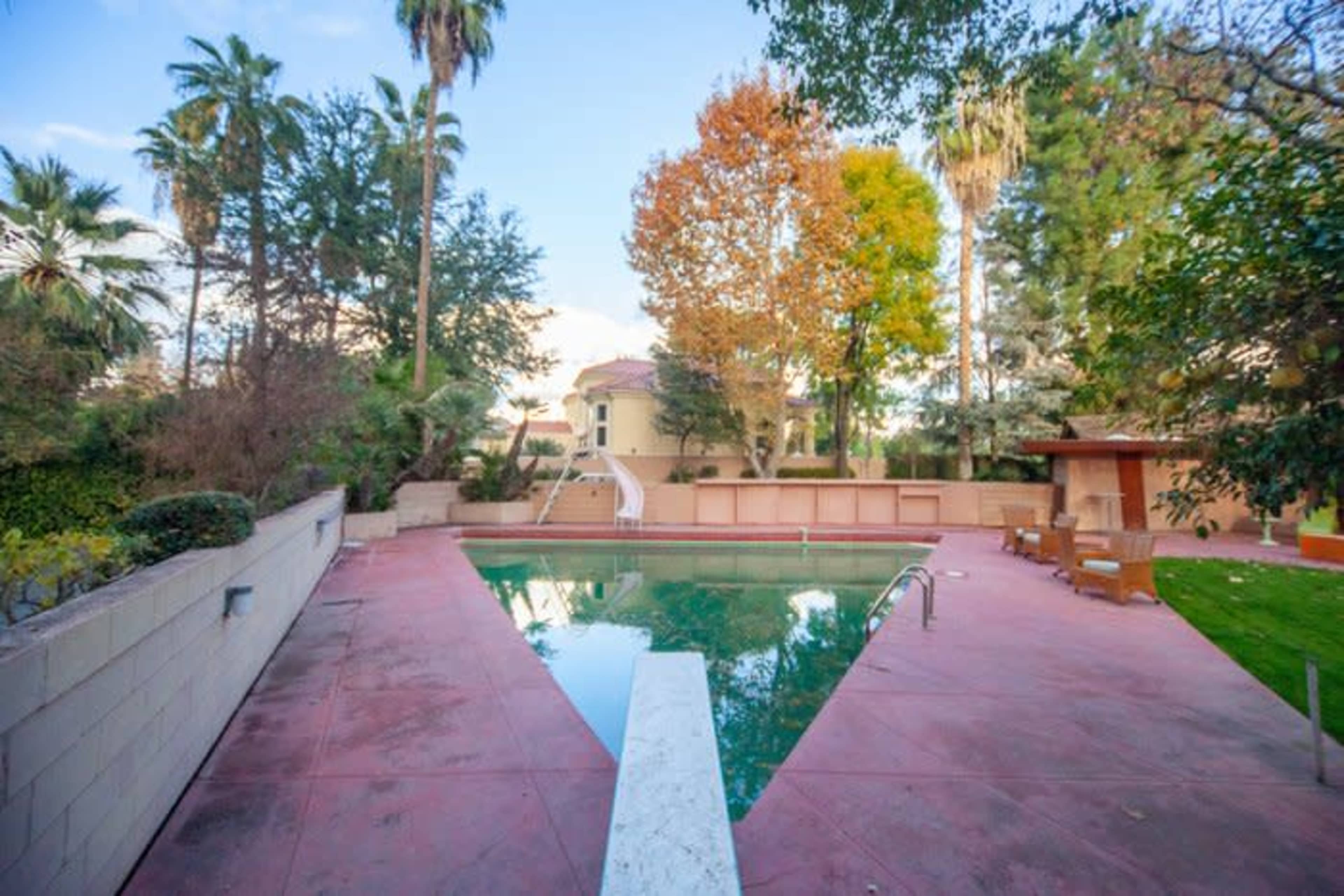 A swimming pool surrounded by palm trees and other greenery, with a large building visible in the background.