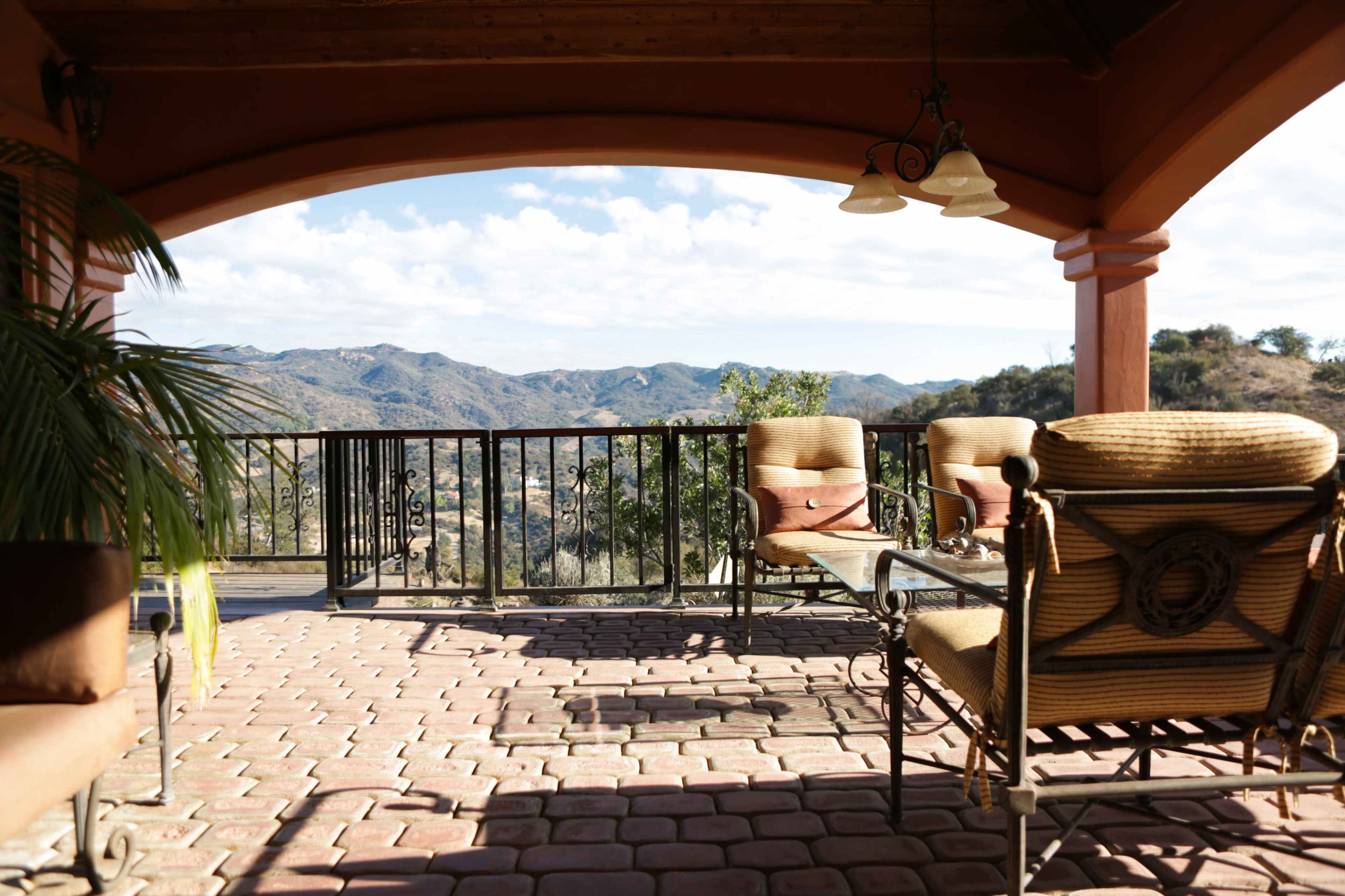 A patio with two chairs and a table overlooks a mountainous landscape, framed by an archway and black railings.