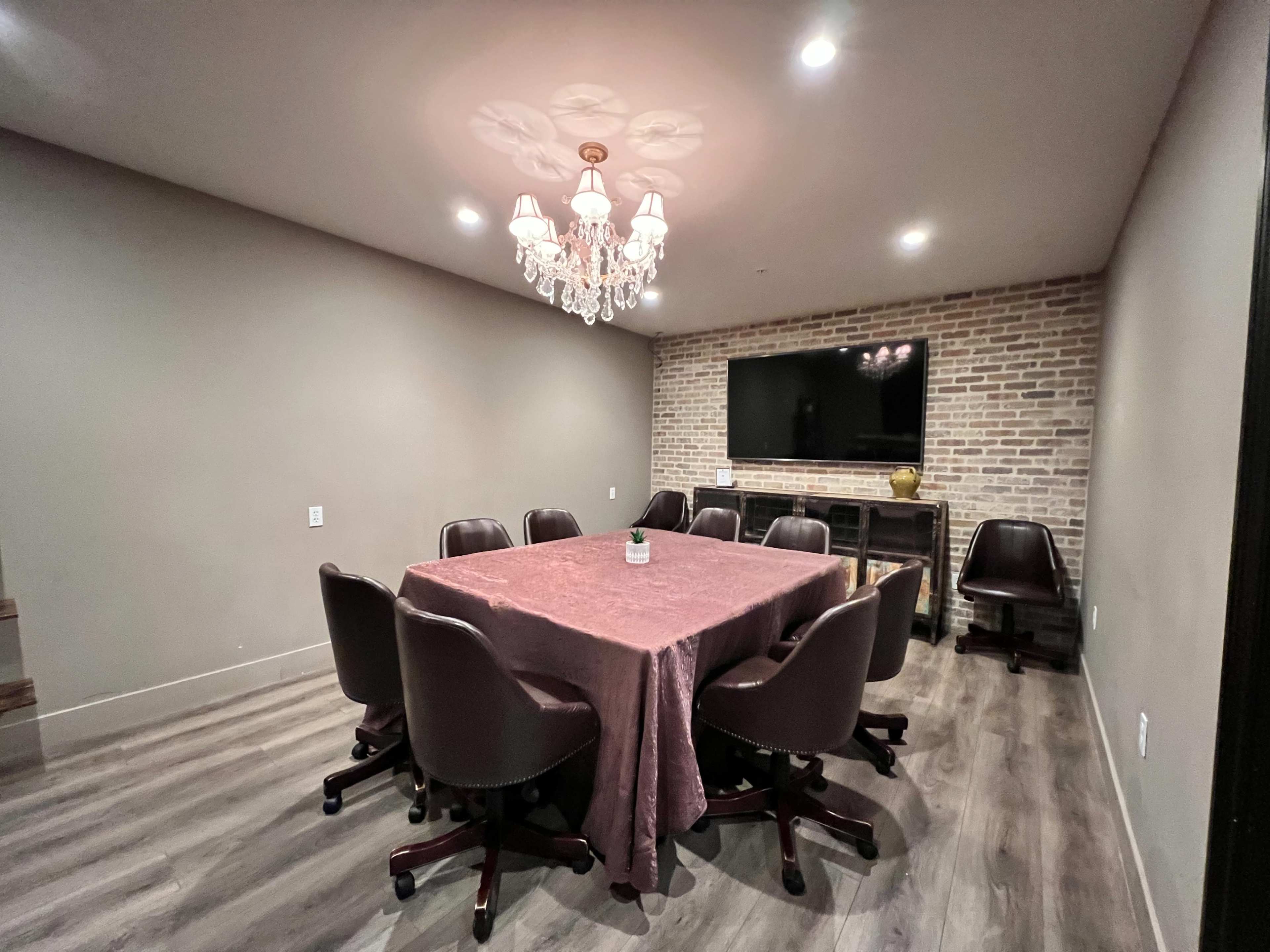 A conference room with a large table surrounded by eight brown chairs, a chandelier above, and a brick wall with a television mounted on it.