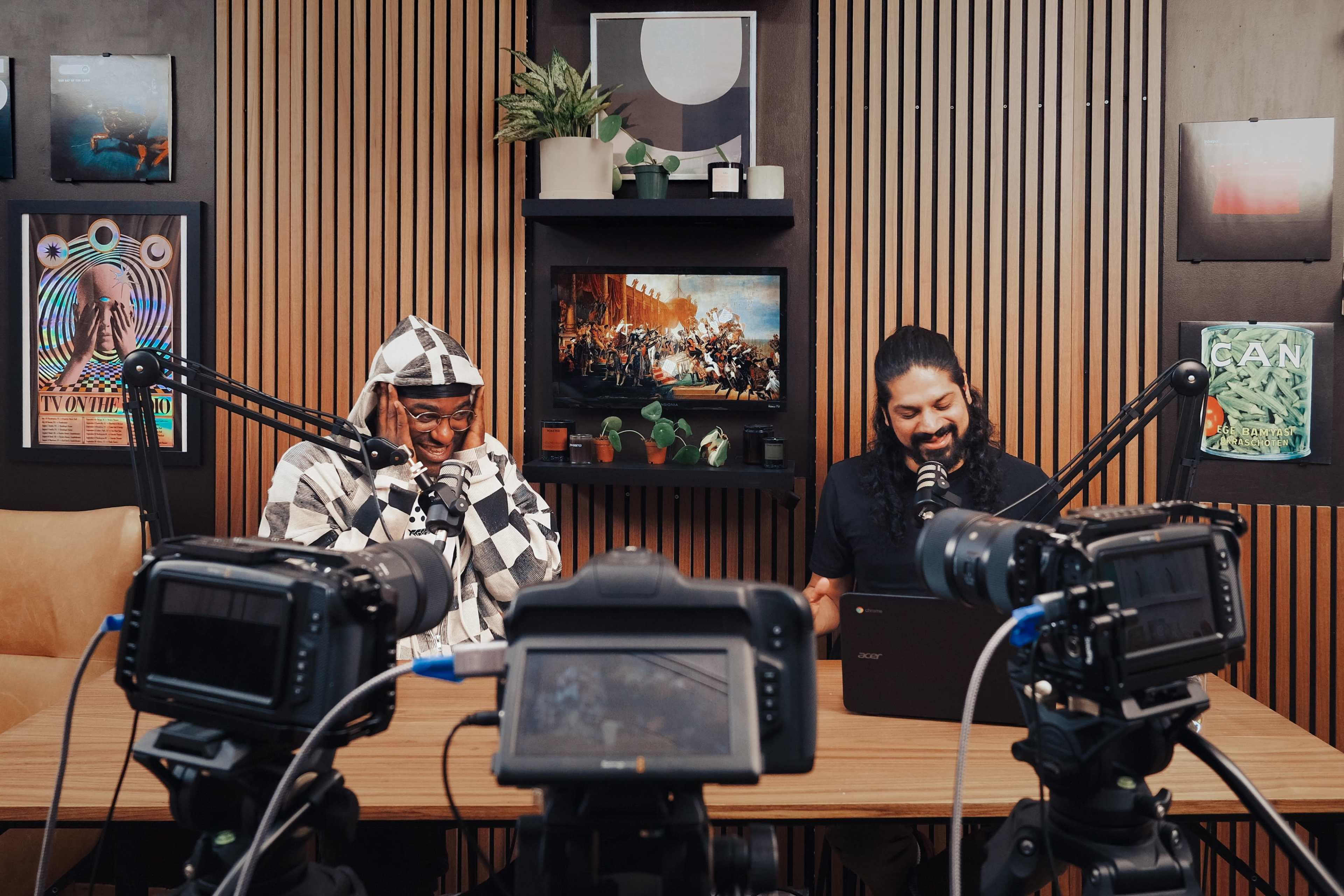 Two men are seated at a table in a podcast studio, surrounded by multiple cameras and framed artwork, while discussing content displayed on a screen behind them.
