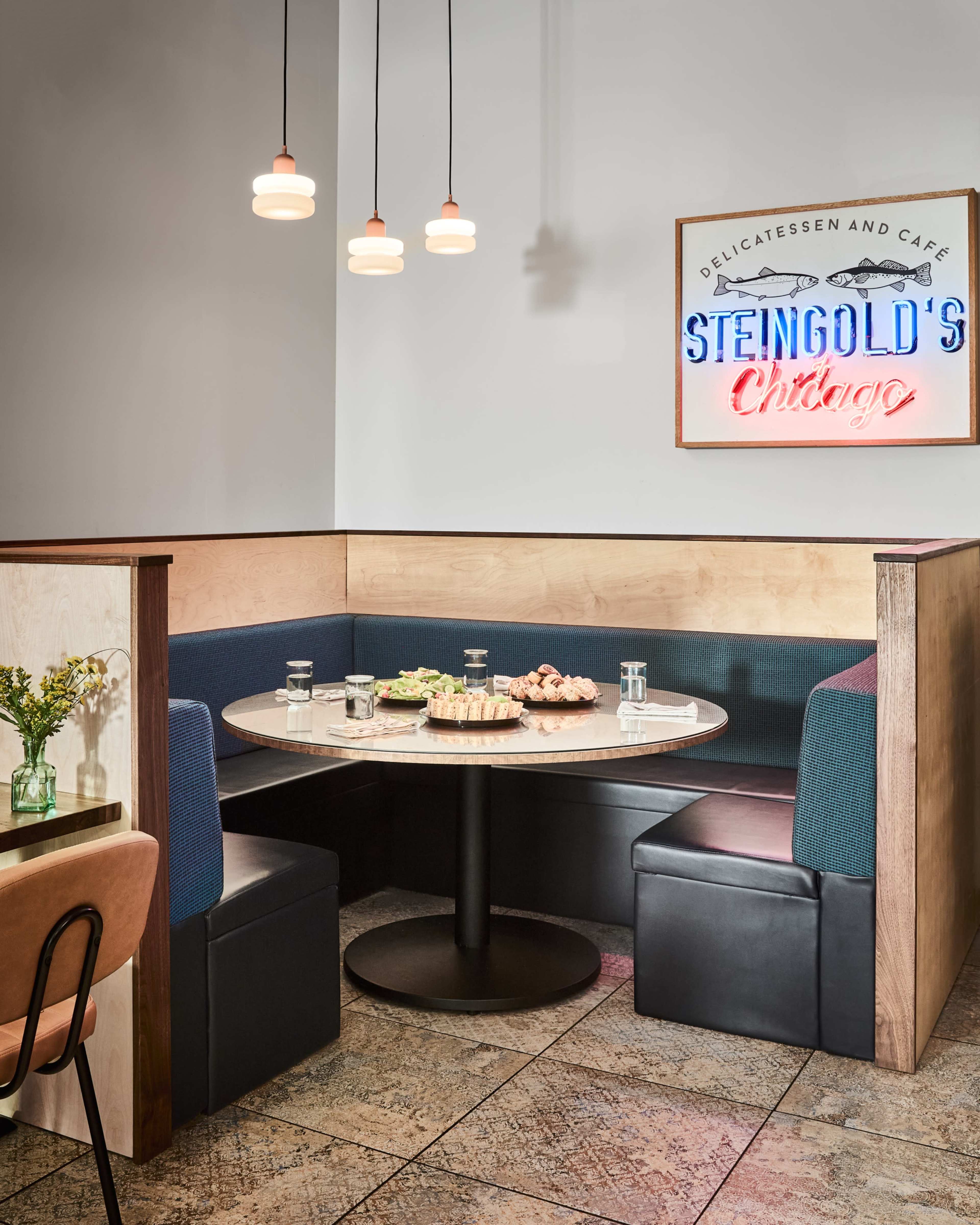A corner booth in a cafe features a round table with a variety of food items and drinks, surrounded by blue and black seating, under a neon sign that reads "STENGOLD'S Chicago."