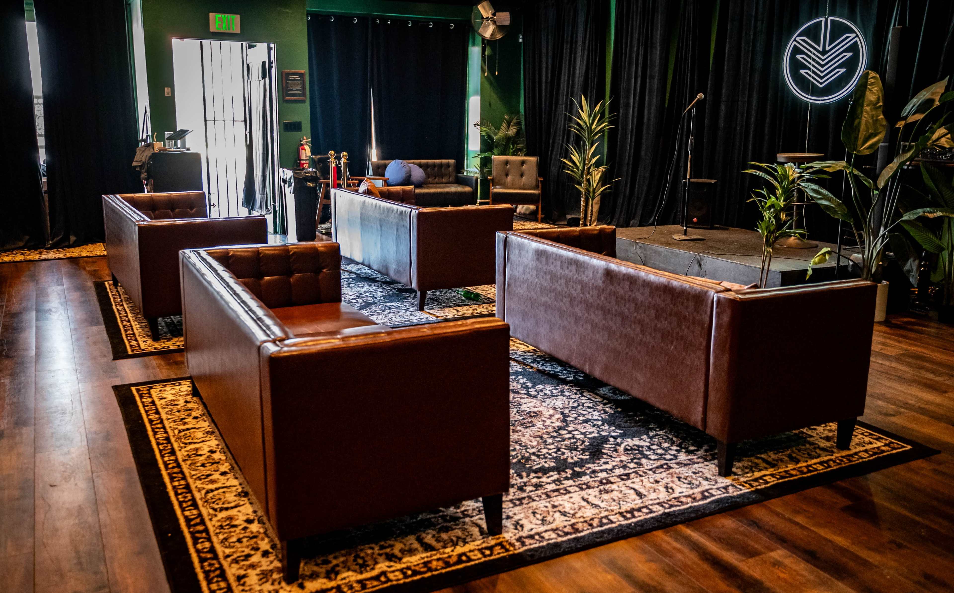 A cozy lounge area with brown leather sofas arranged around a central rug, facing a small stage surrounded by plants.
