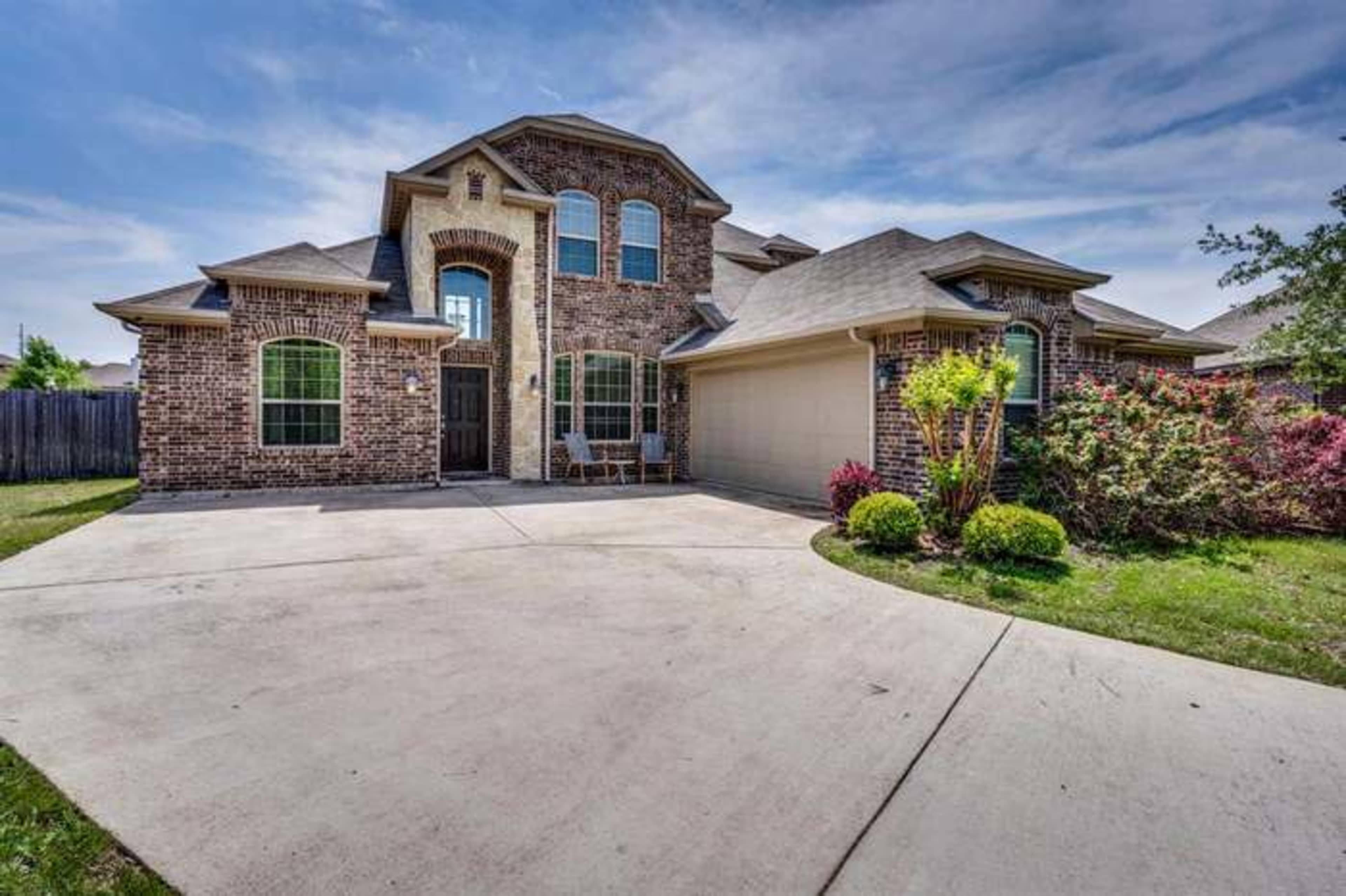 The image shows a two-story brick home with a concrete driveway, large windows, and a landscaped front yard.
