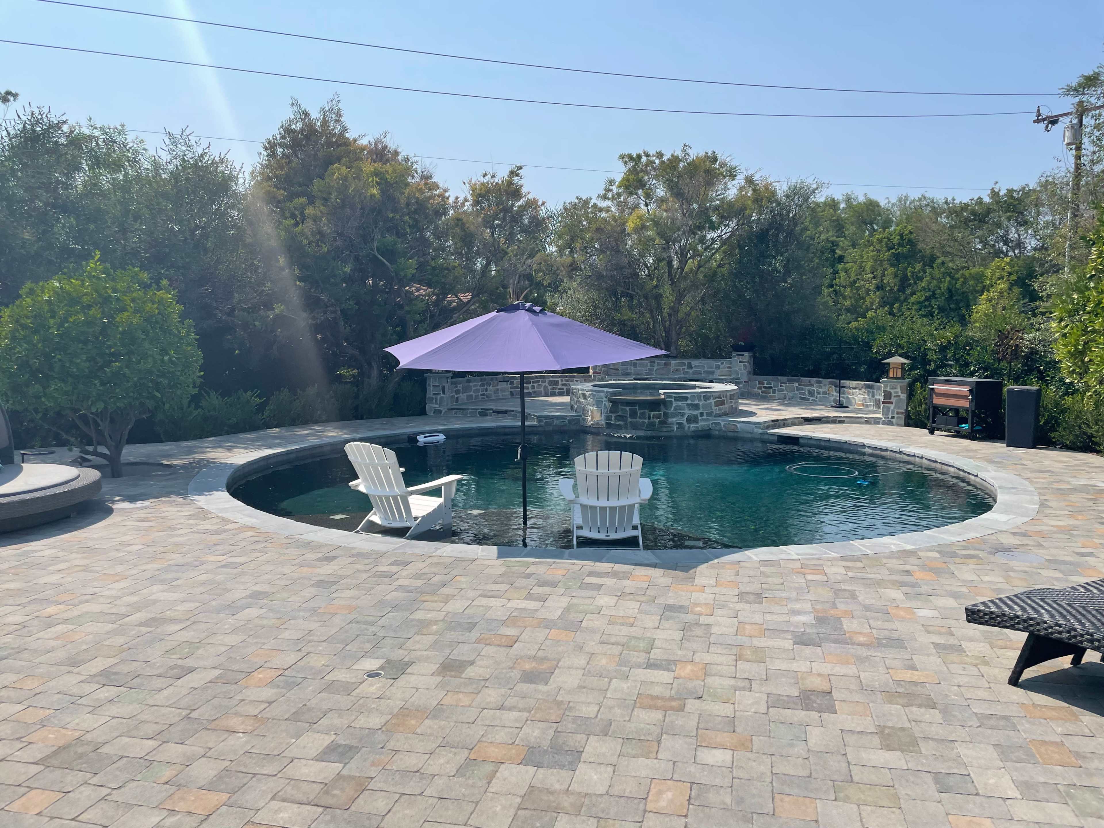 The image shows a circular pool surrounded by a paved patio with two white chairs under a purple umbrella and lush greenery in the background.